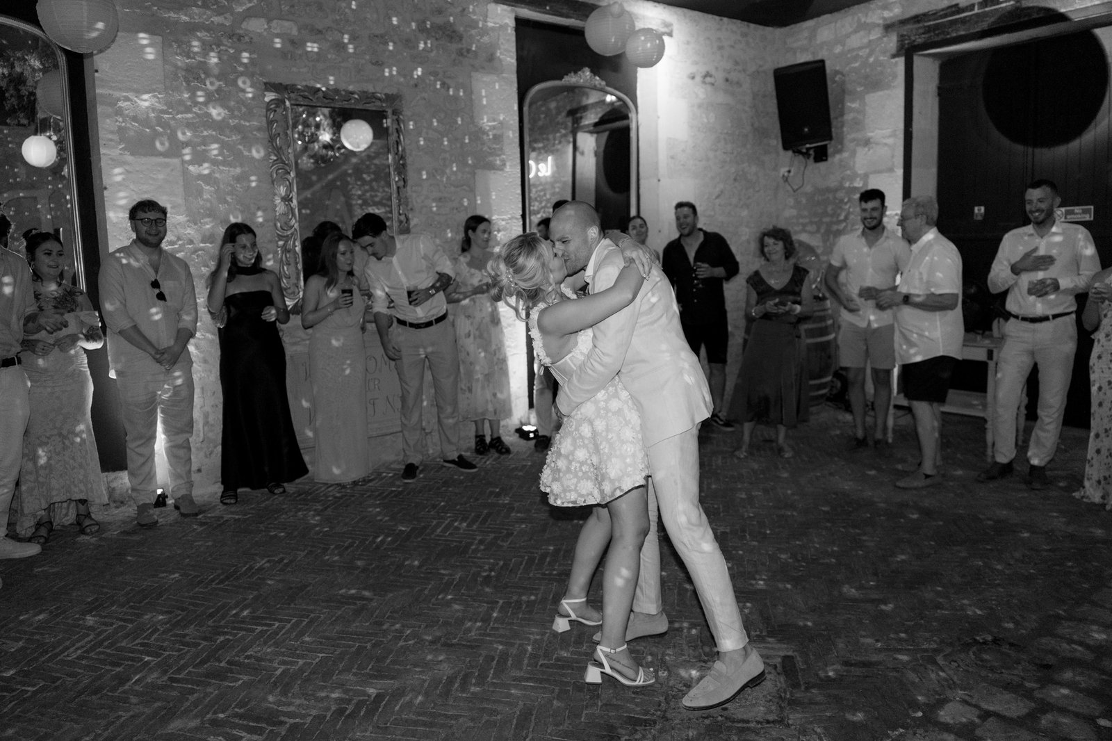 A couple embraces and dances in the center of a room while guests stand around them, watching and clapping. The scene appears to be a celebration or party in a stone-walled venue.