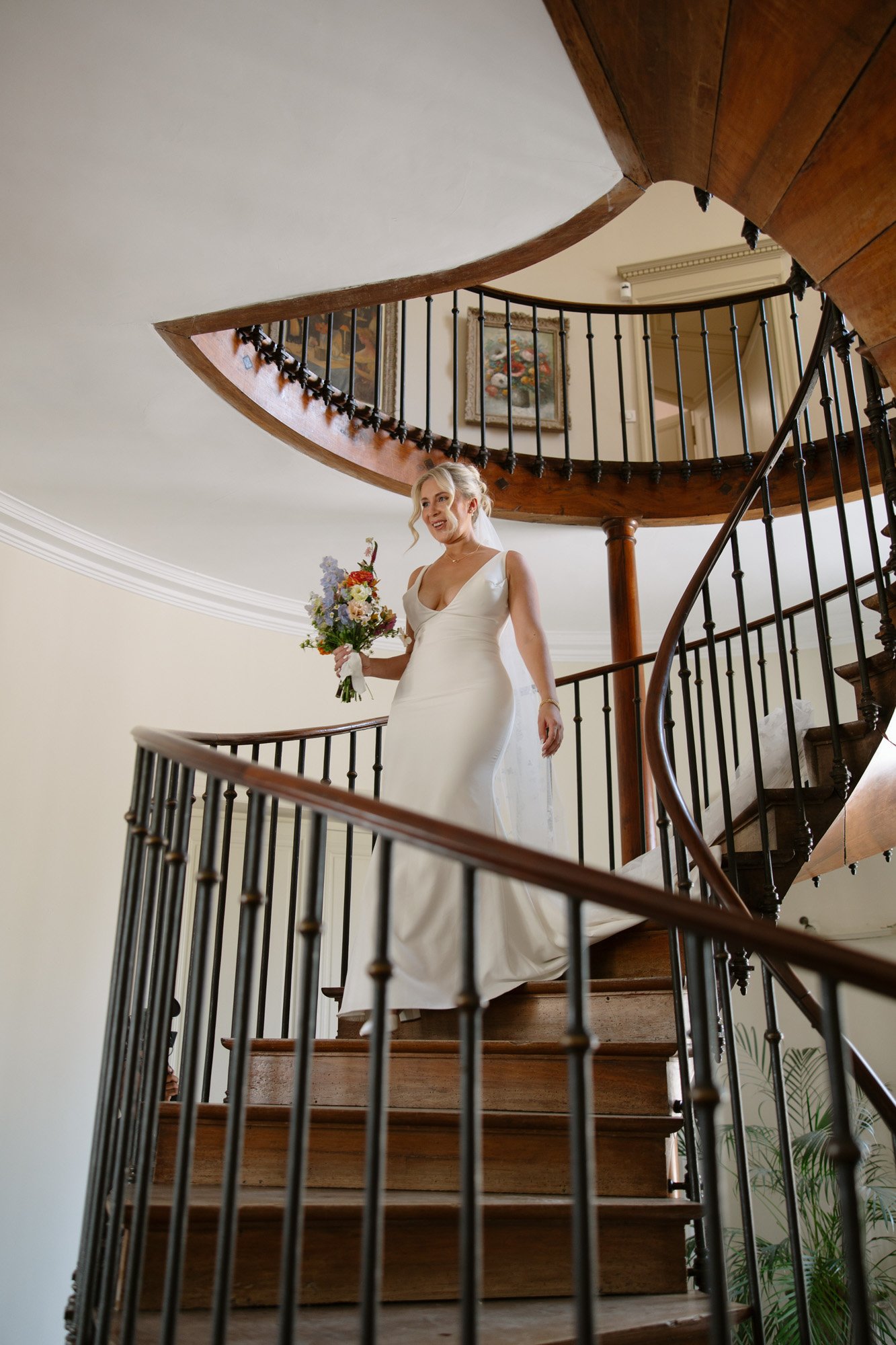 A woman in a white wedding dress holding a bouquet descends a curved wooden staircase with black railings.
