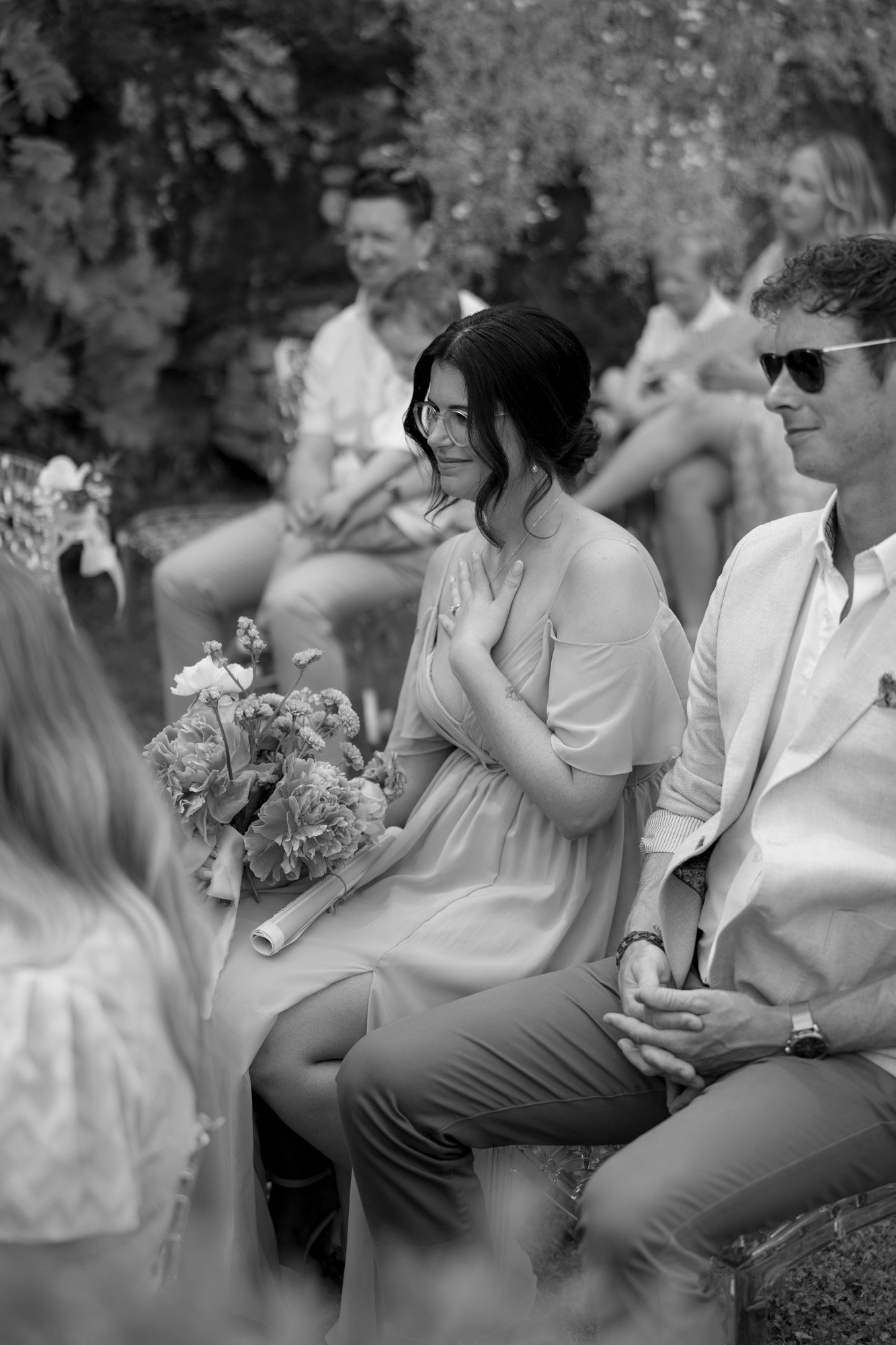 A woman in a light dress smiles while seated outdoors among other guests at an event, holding a bouquet of flowers.