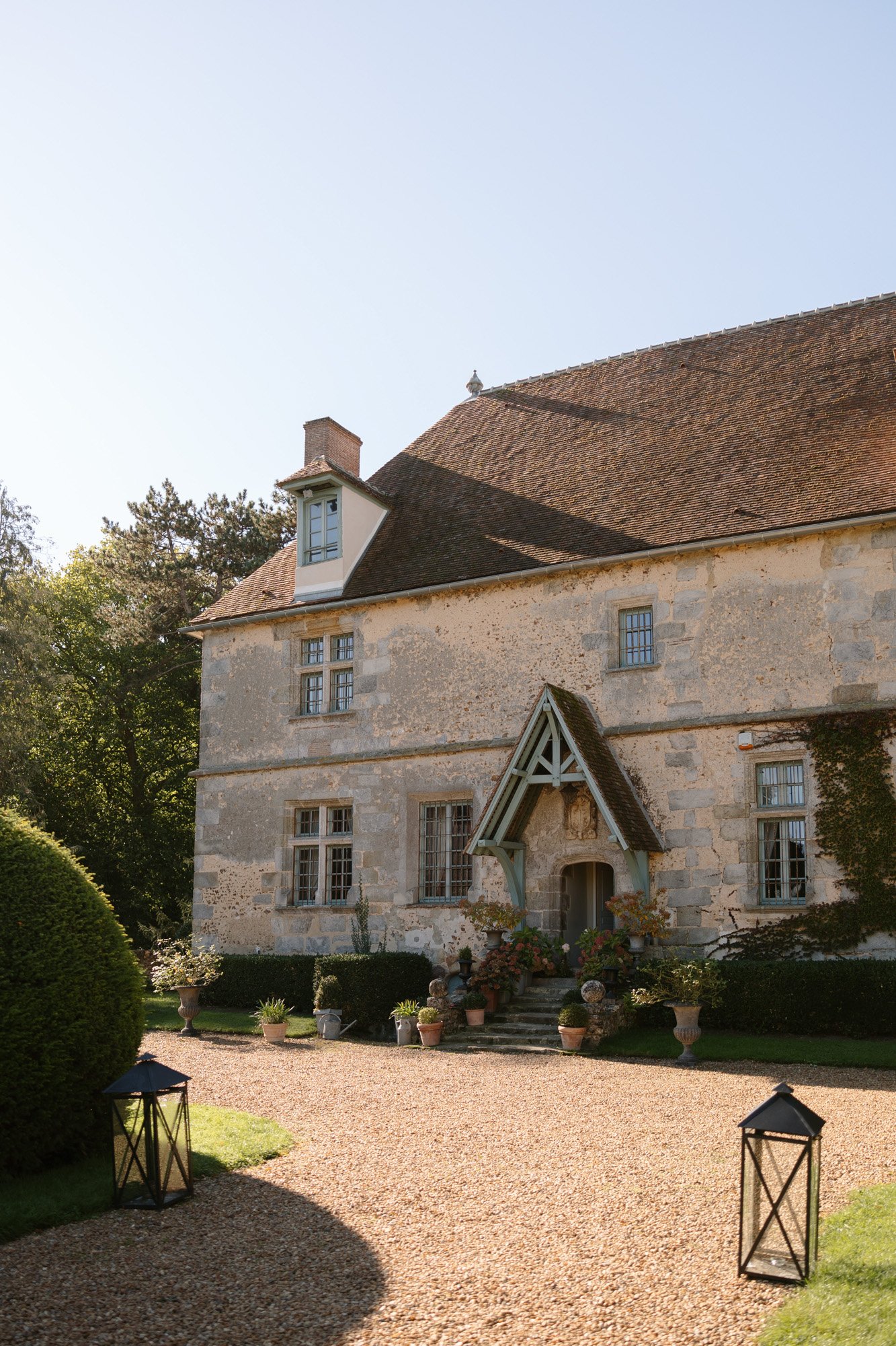 A stone country house with a steep roof, potted plants at the entrance, a gravel path, and black lanterns on the lawn. Manoir de Vacheresses wedding.