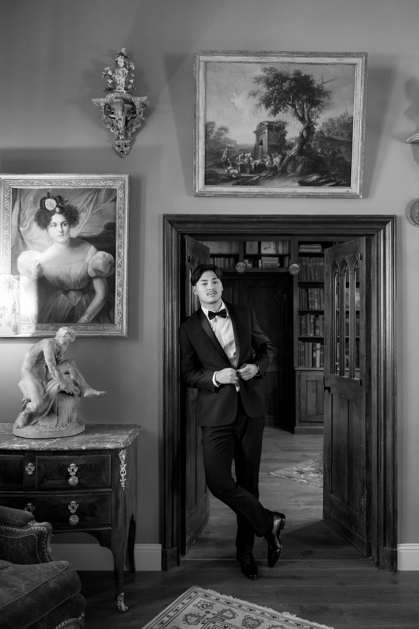 A man in a tuxedo stands in a doorway of an elegant room decorated with paintings, sculptures, and ornate furnishings. Manoir de Vacheresses wedding.