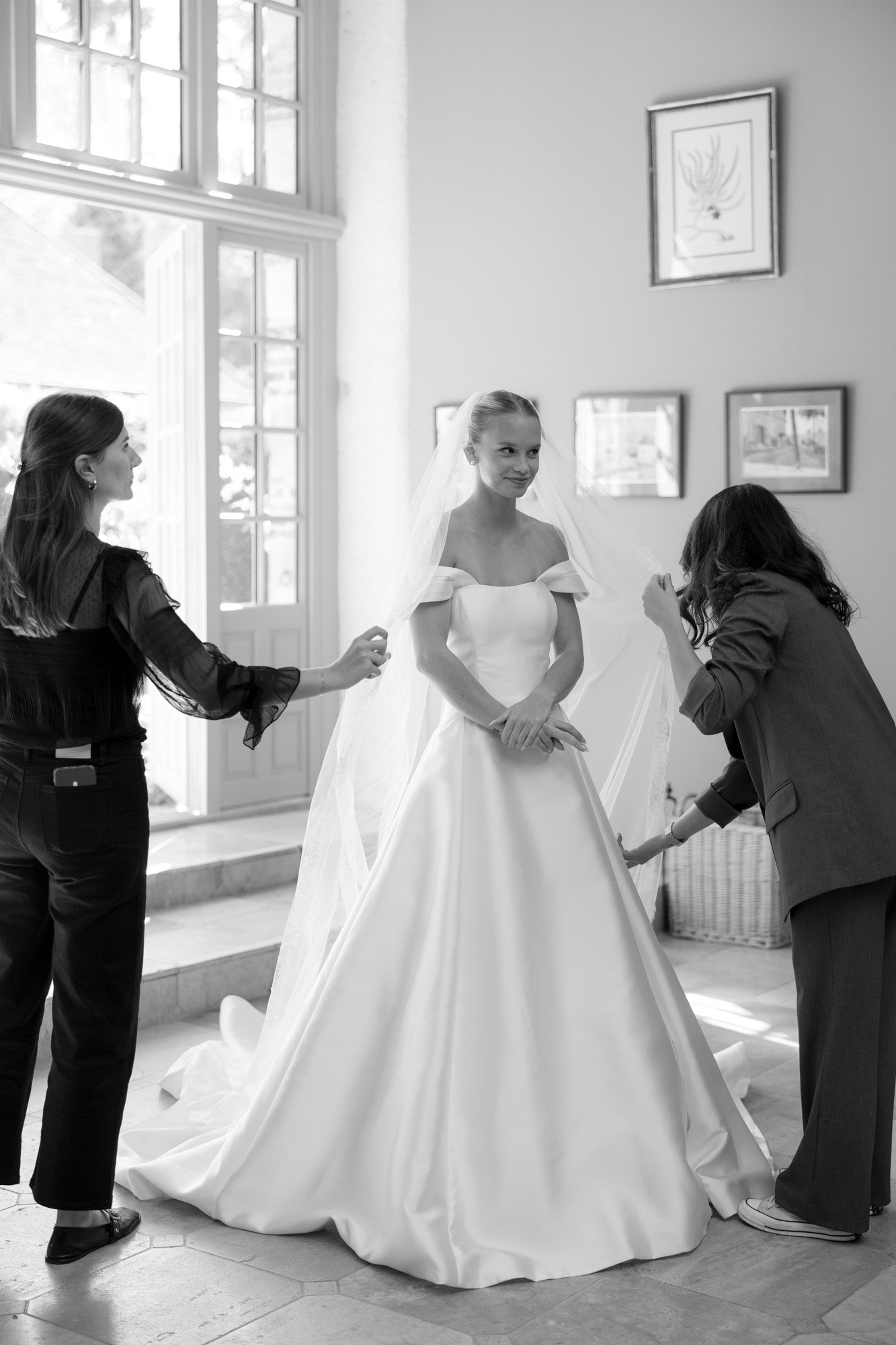 A bride in a white gown stands indoors while two women adjust her veil and dress. Framed pictures hang on the wall behind them.