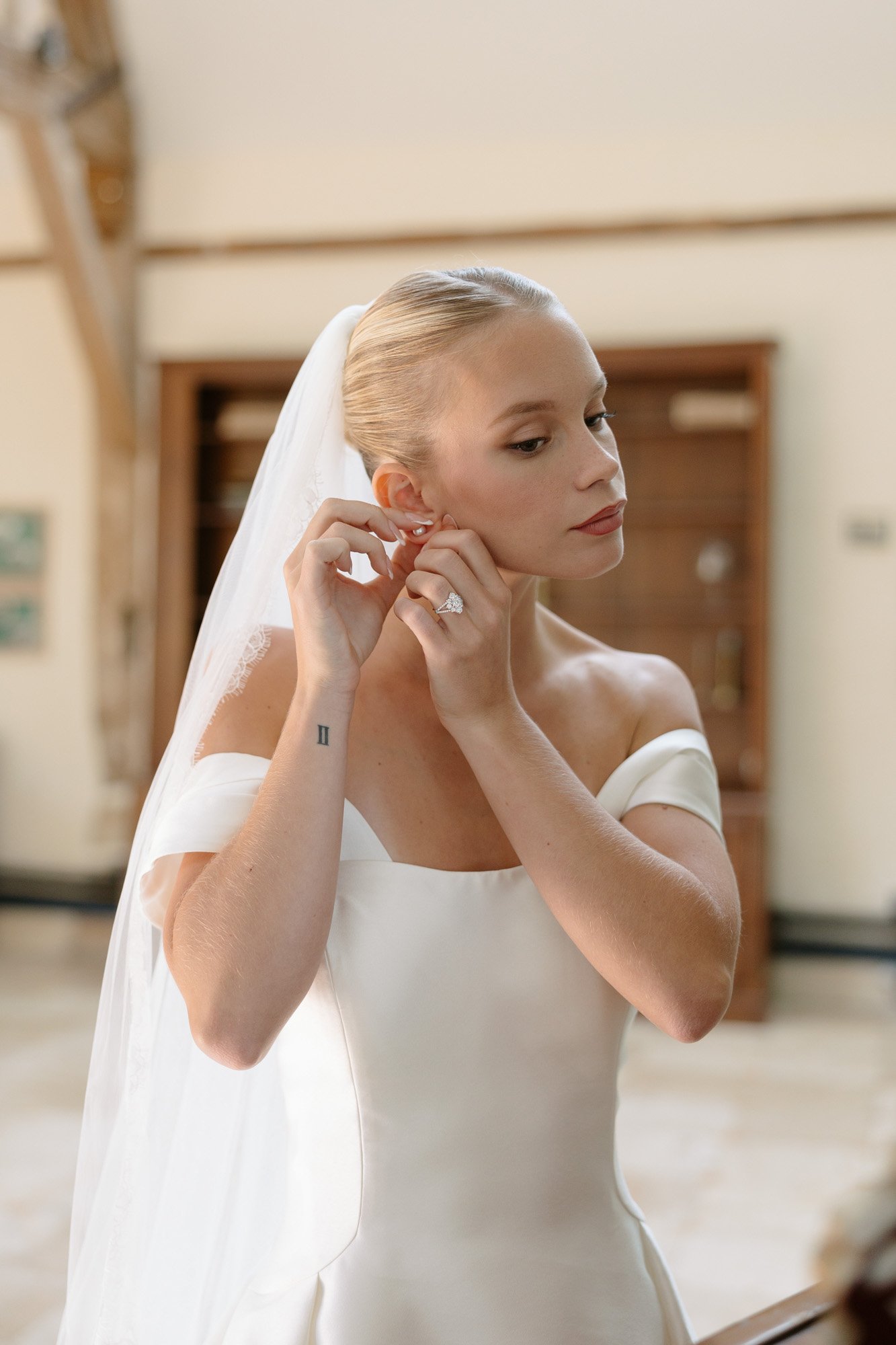 A bride in an off-the-shoulder white gown and veil puts on an earring while standing indoors.
