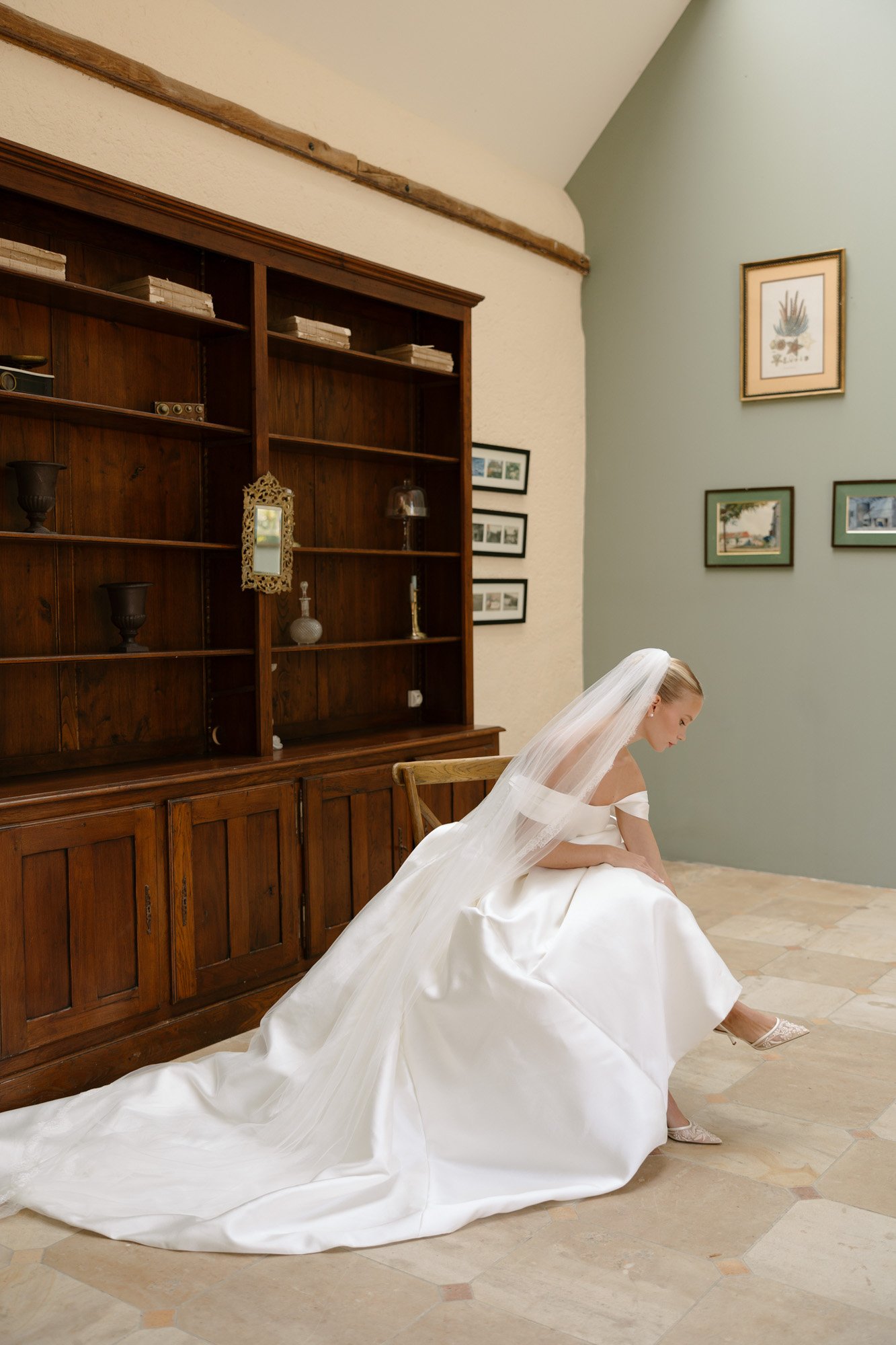 A bride in a white gown and veil sits on a chair adjusting her shoe in a room with a wooden cabinet and framed pictures on the wall. Manoir de Vacheresses wedding.