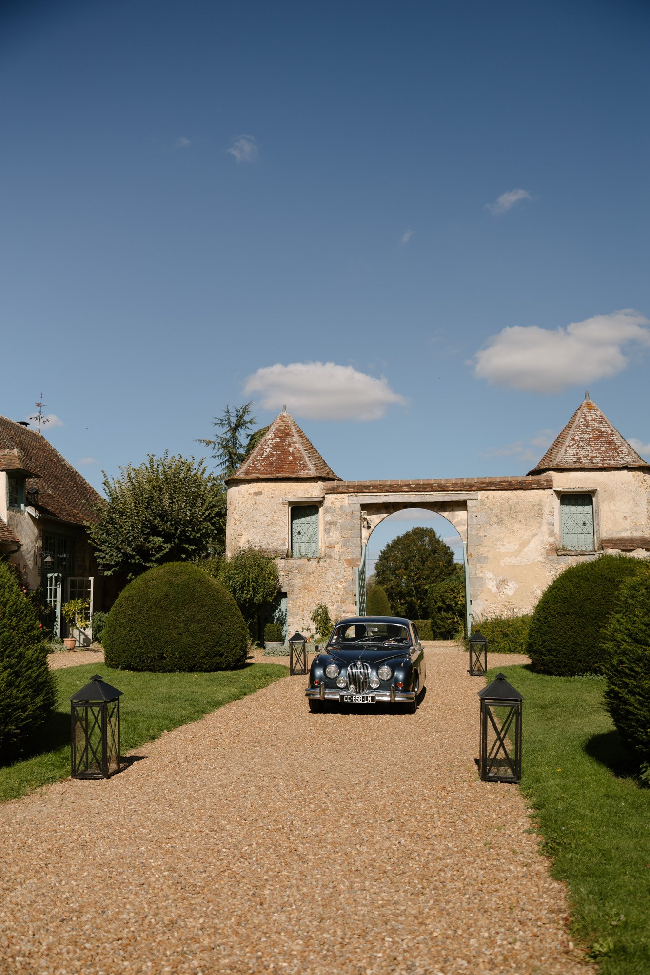A classic black car drives along a gravel path toward a stone archway, flanked by trimmed bushes and lanterns, under a clear blue sky. Manoir de Vacheresses wedding.
