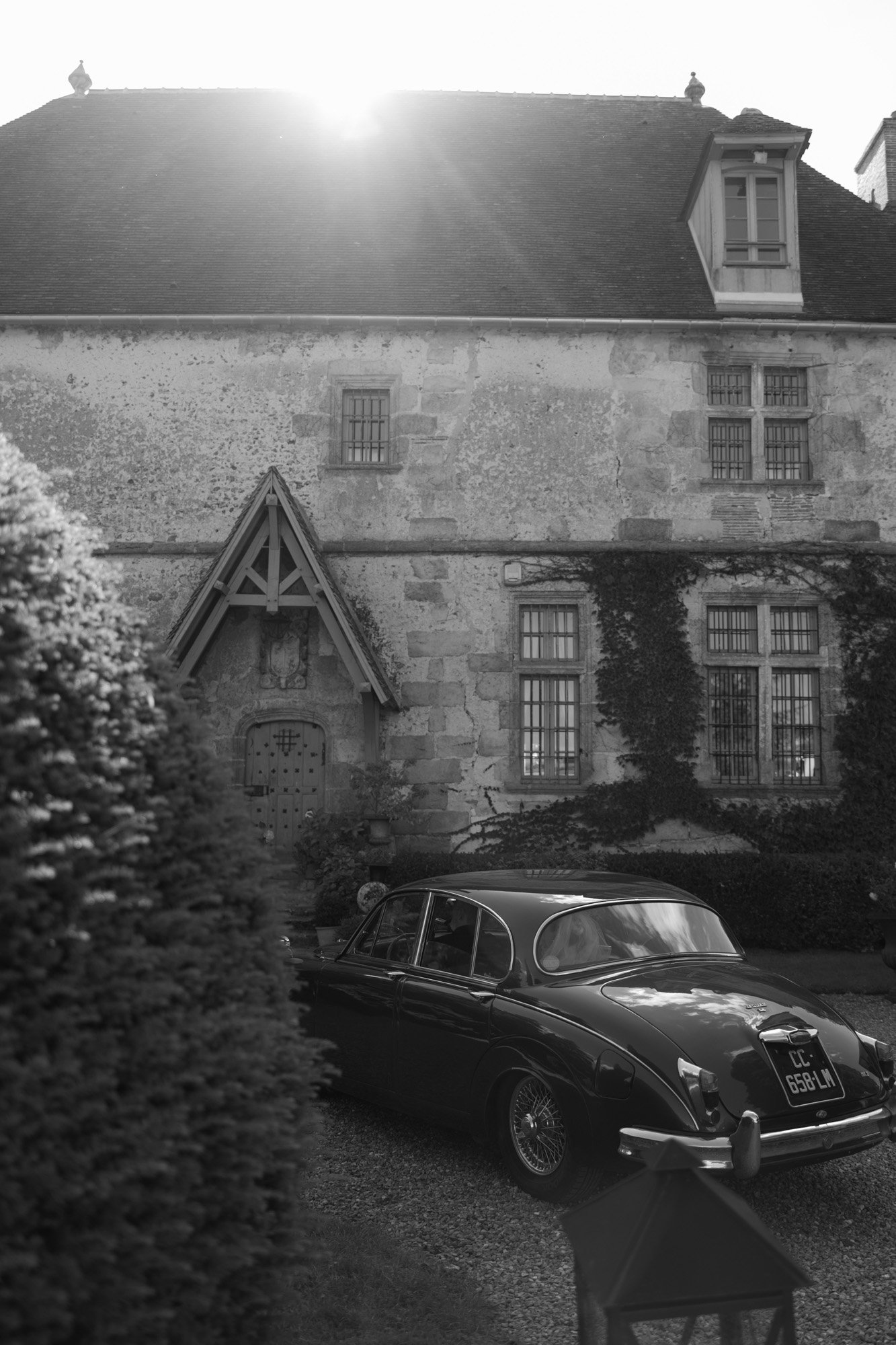 A vintage car is parked in front of an old stone house with tall windows and ivy on the walls, as sunlight shines from behind the roof.