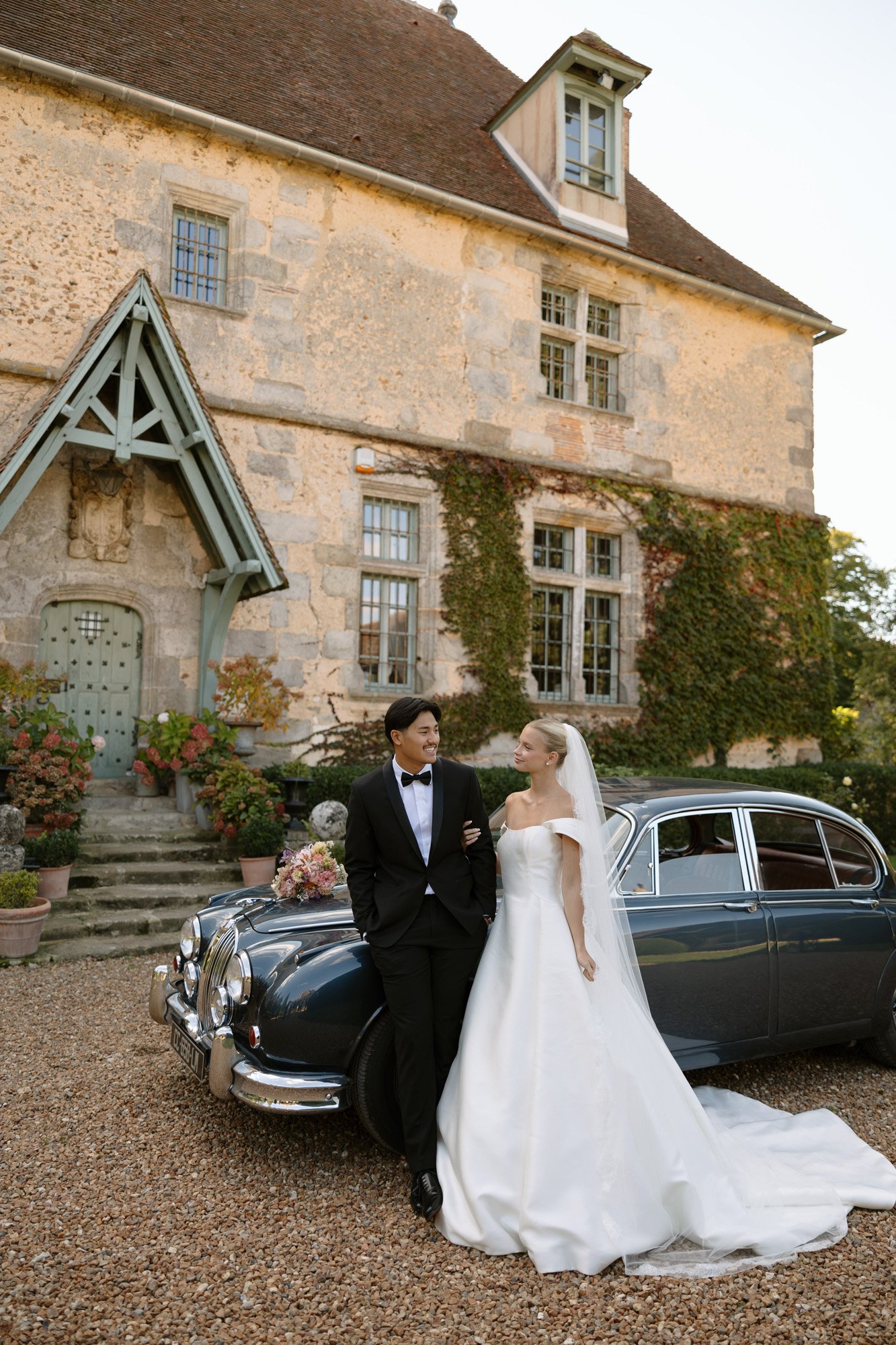 A bride and groom stand beside a vintage car in front of a historic stone building covered with ivy. Manoir de Vacheresses wedding.