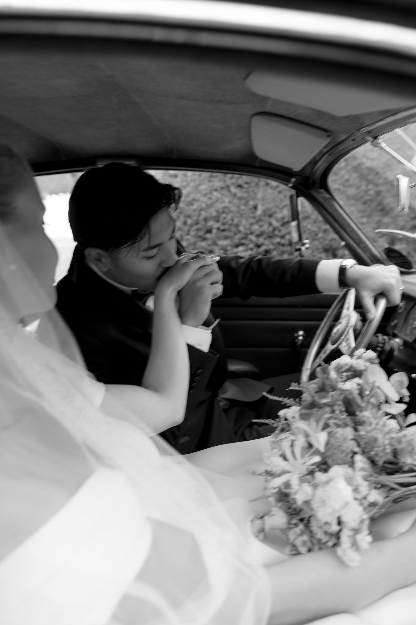 A groom kisses the bride’s hand while sitting together in the front seat of a vintage car; the bride holds a bouquet.