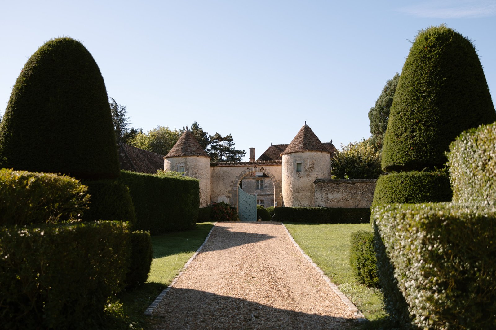 Gravel pathway lined with trimmed hedges leads to a stone building with two round towers and a blue gate under a clear sky.
