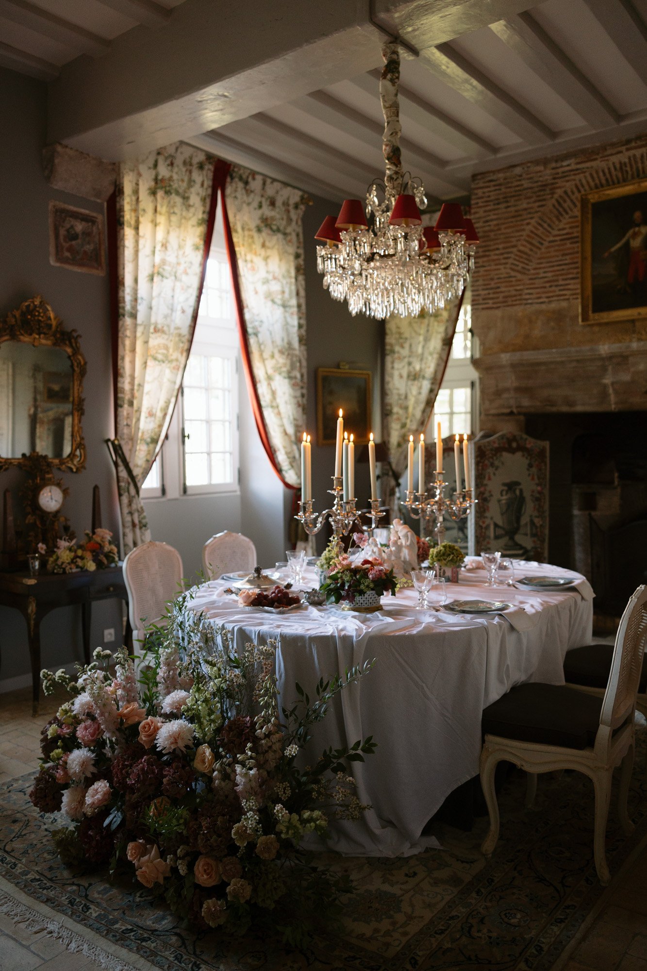 Elegant dining room with a round table set for a meal, draped white tablecloth, tall candles, floral arrangements, chandelier, and ornate decor in a softly lit, classic interior. Manoir de Vacheresses wedding.