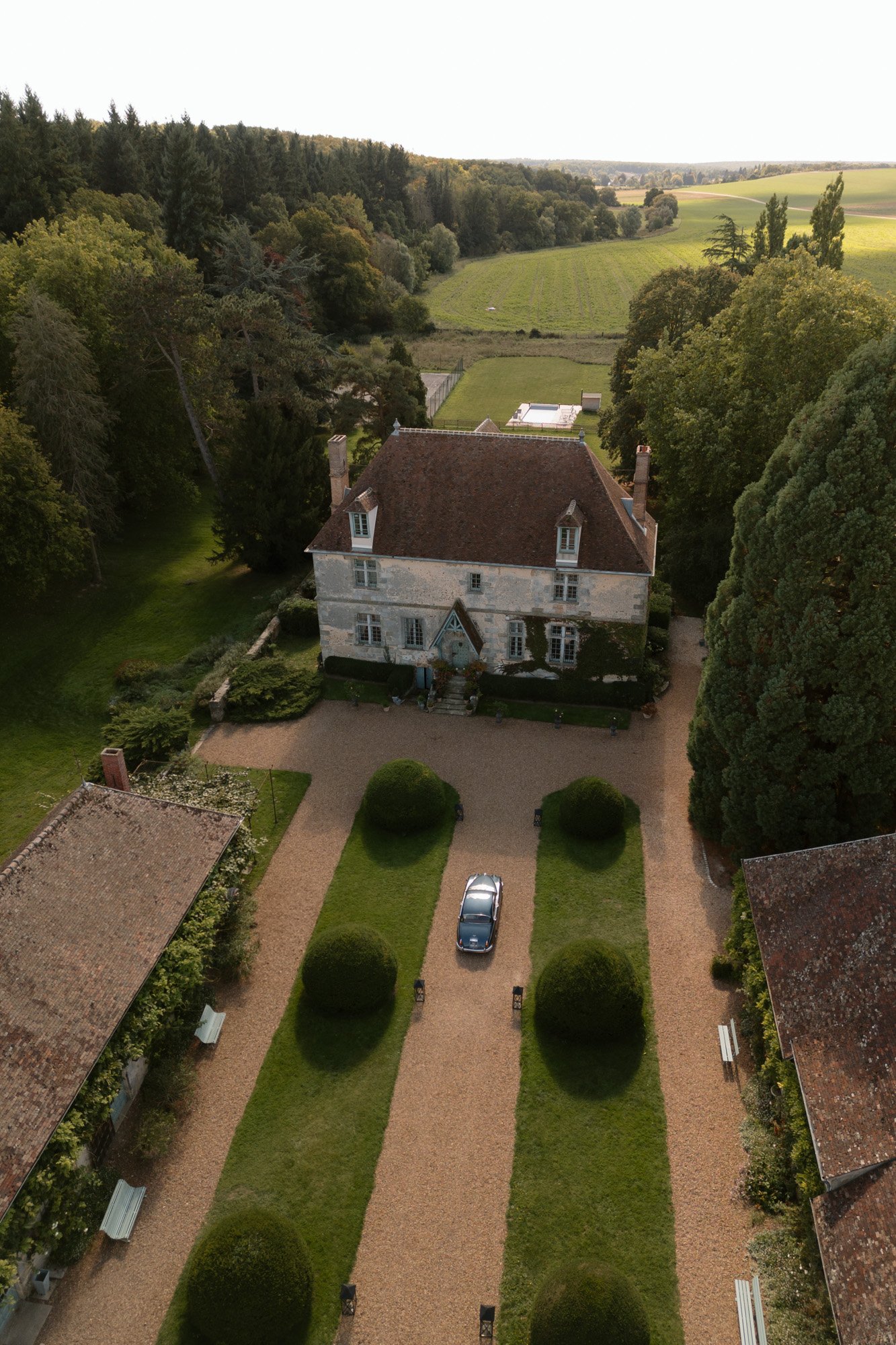 Aerial view of a country house with a red roof, surrounded by trees and gardens, with a car parked on the gravel driveway in front. Manoir de Vacheresses wedding.
