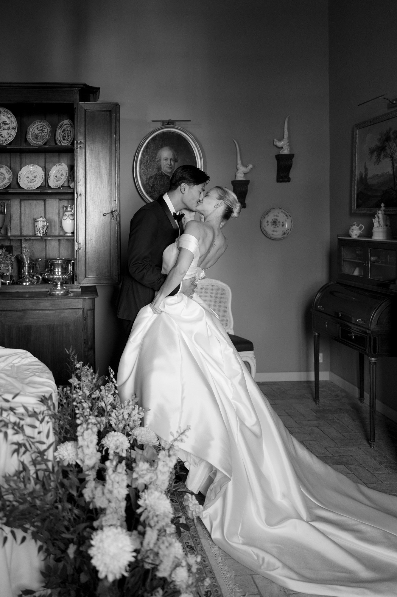 A bride and groom kiss indoors next to a cabinet and a desk; the bride wears an off-shoulder gown with a long train, and the groom is in a tuxedo. Manoir de Vacheresses wedding.