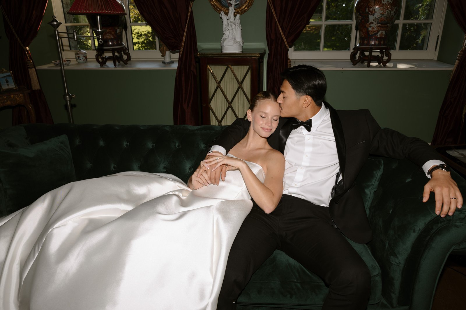 A couple dressed in formal wedding attire sits on a green velvet couch; the woman rests against the man, who kisses her on the forehead.  Manoir de Vacheresses wedding.