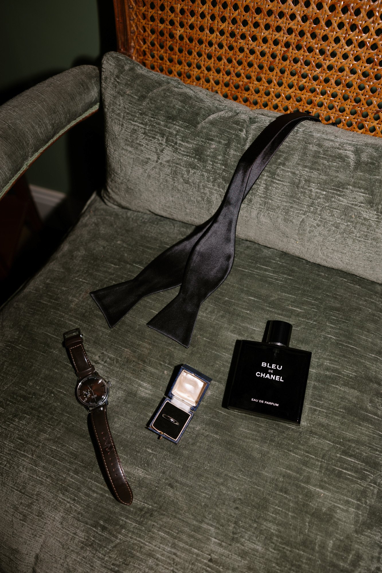 A black bow tie, a bottle of Bleu de Chanel perfume, a wristwatch, and a ring in a box are arranged on a green velvet chair.