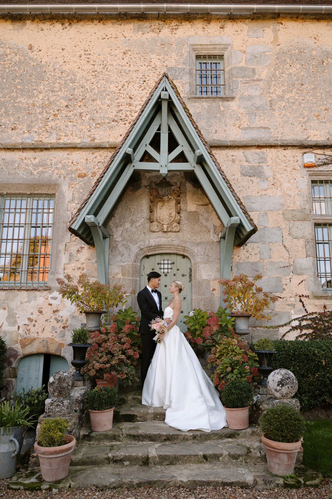 A bride and groom in formal attire stand on stone steps in front of a rustic building with potted plants and a wooden awning.