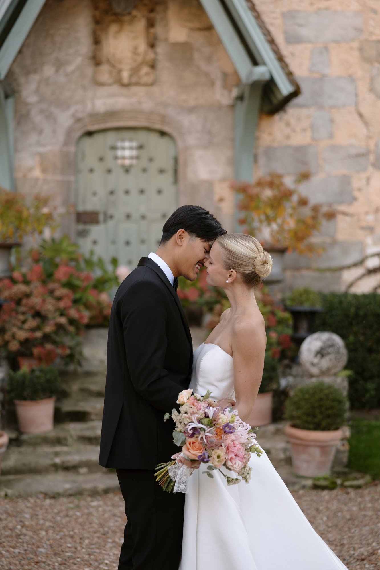 A bride and groom stand close together outside a stone building, touching foreheads and holding a bouquet of flowers. Manoir de Vacheresses wedding.