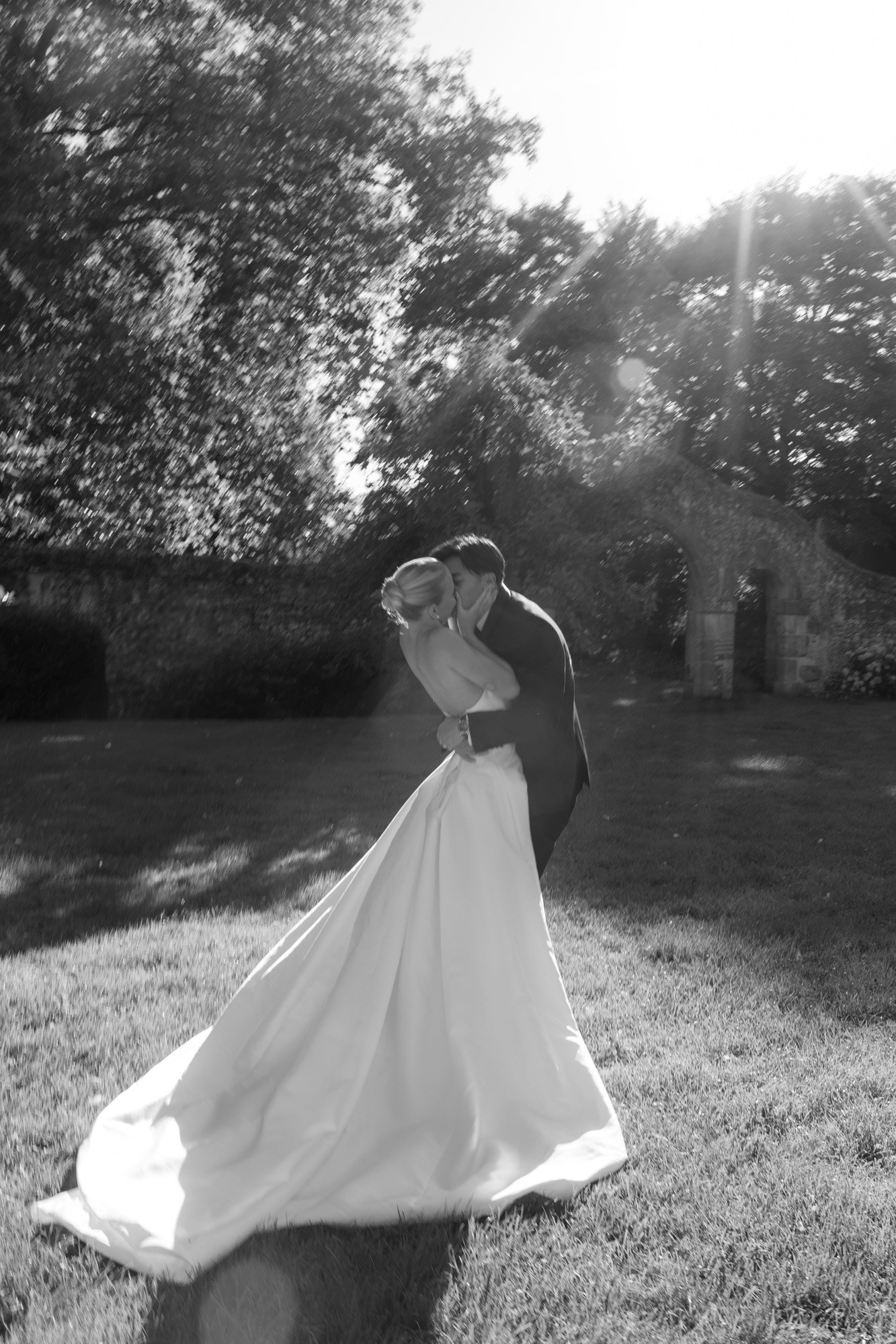 A bride and groom share a kiss outdoors in sunlight, with trees and an old stone wall in the background.
