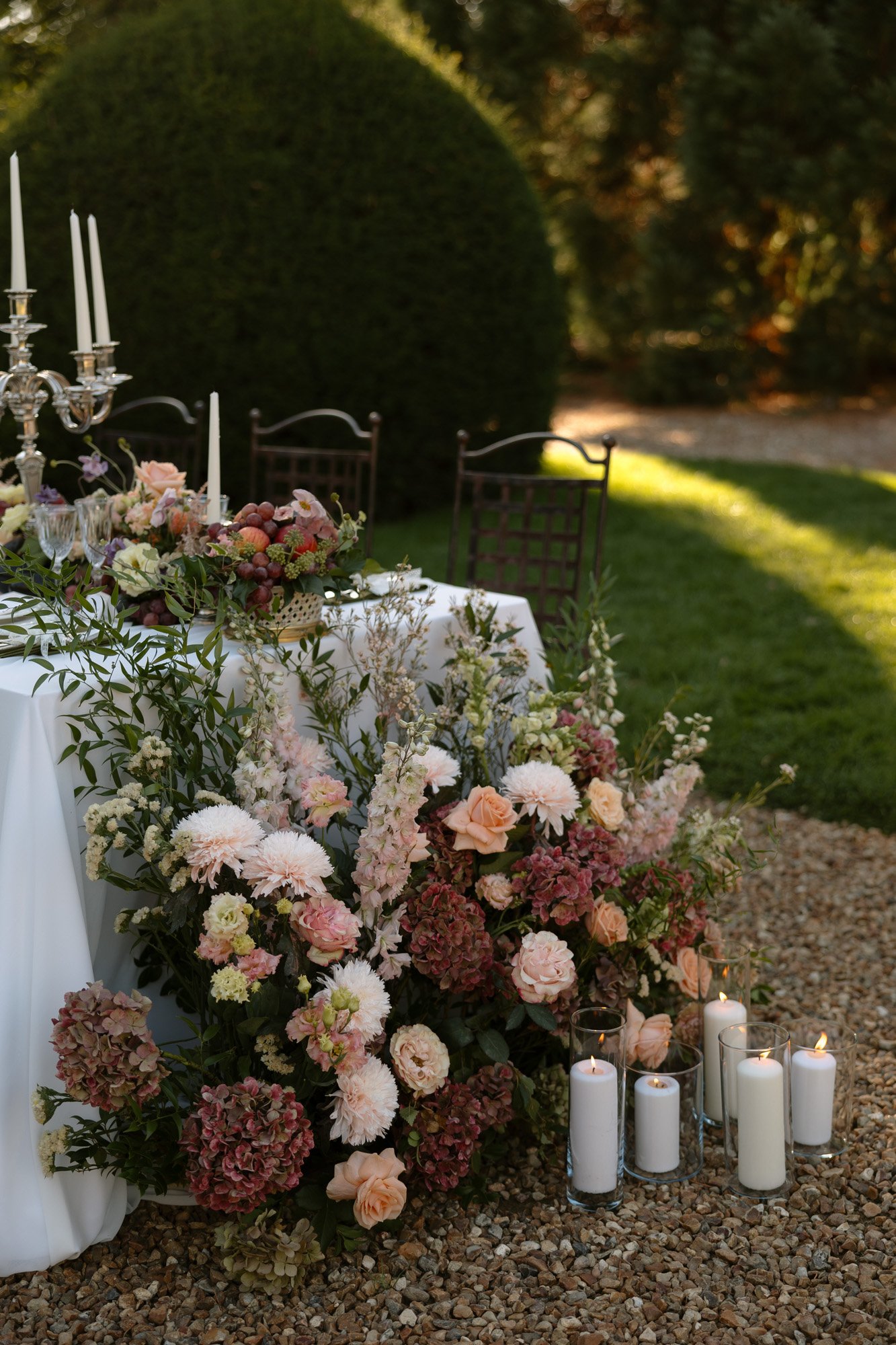 A decorated outdoor table with white linens, floral arrangements in pastel colors, white candles, and two empty chairs on a gravel surface with greenery in the background.