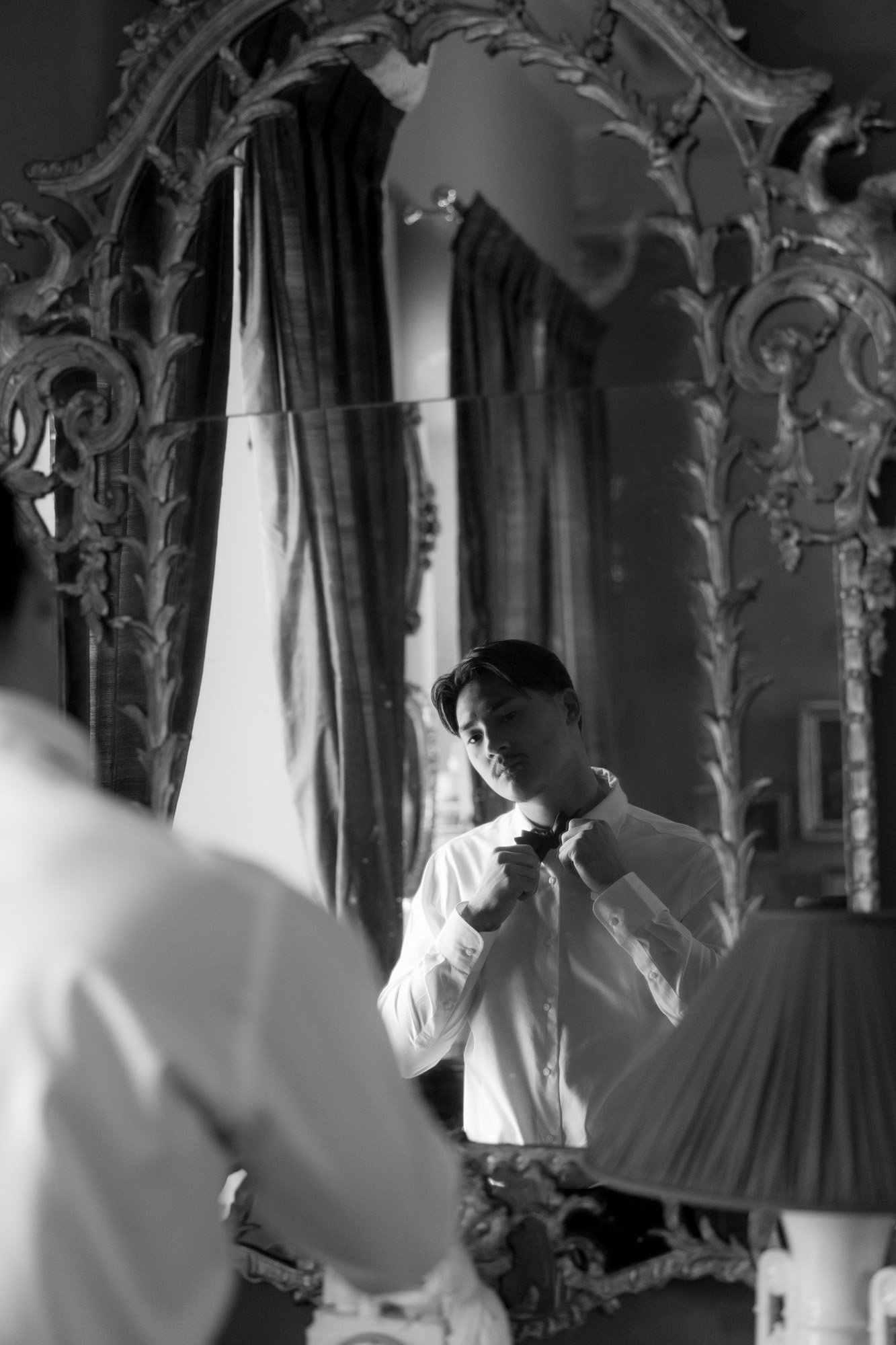 A man in a white shirt is seen in a decorative mirror adjusting his collar in a room with tall curtains and ornate furnishings.