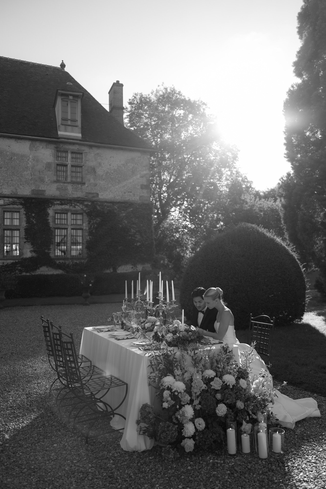 A bride and groom sit together at a flower-adorned outdoor table set for a formal event, with a large house and sunlight in the background. Manoir de Vacheresses wedding.