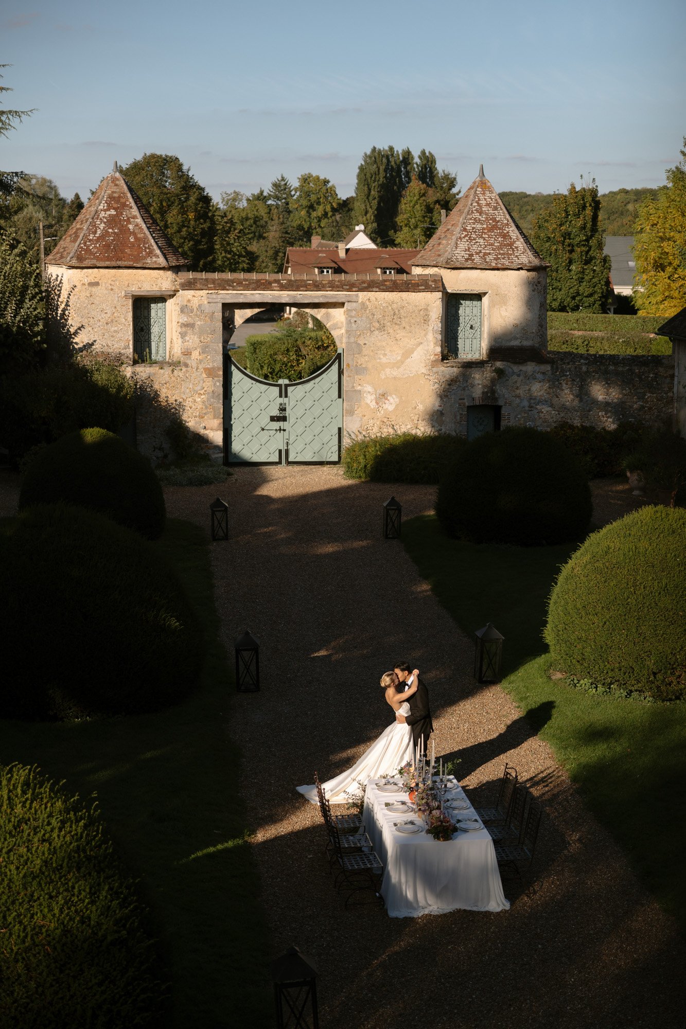 A bride and groom embrace next to a long outdoor dining table set for a meal, with a historic stone gate and gardens in the background under daylight. Manoir de Vacheresses wedding.