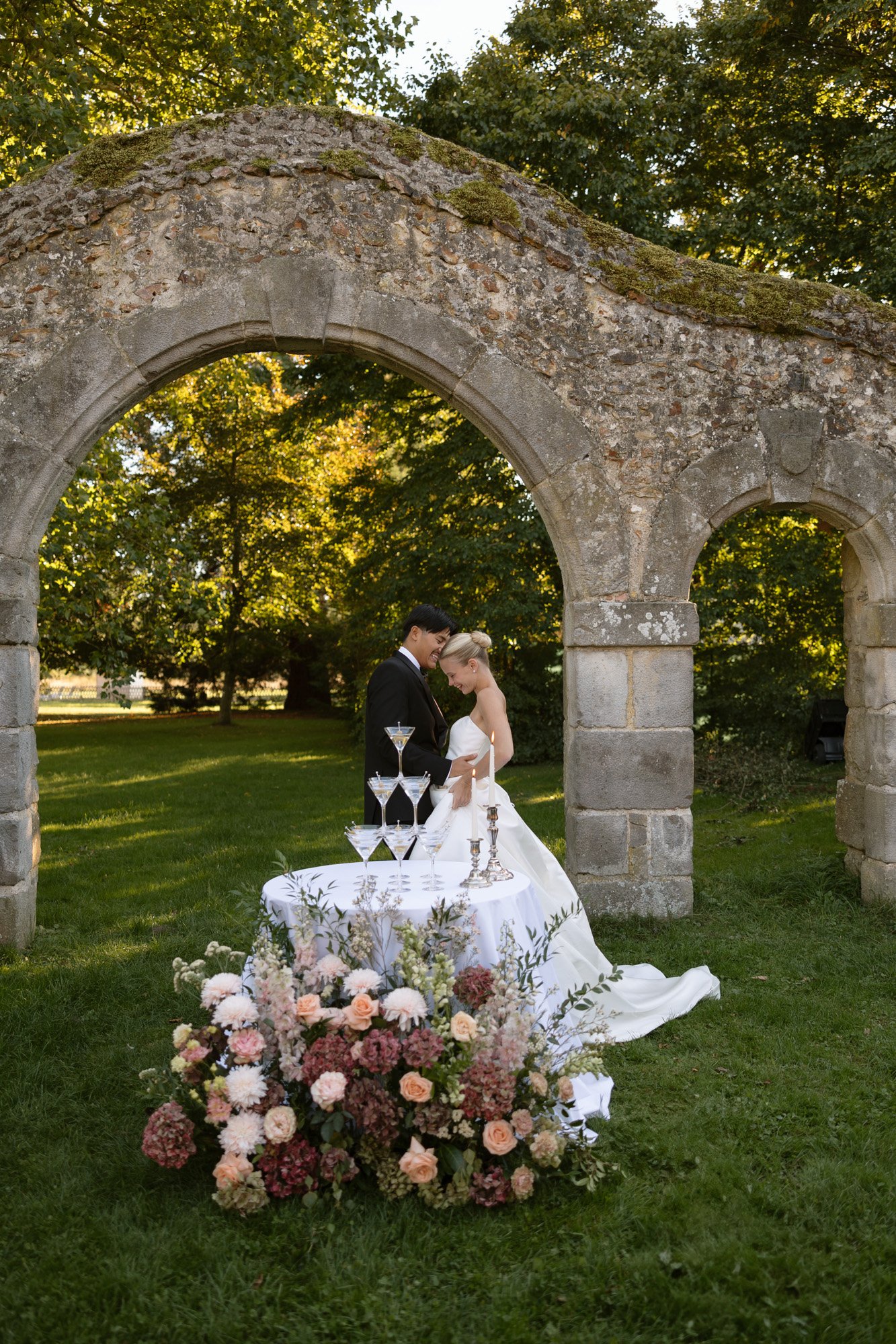 A bride and groom stand close together under a stone arch outdoors, with a decorated table and floral arrangement in front of them. Manoir de Vacheresses wedding.