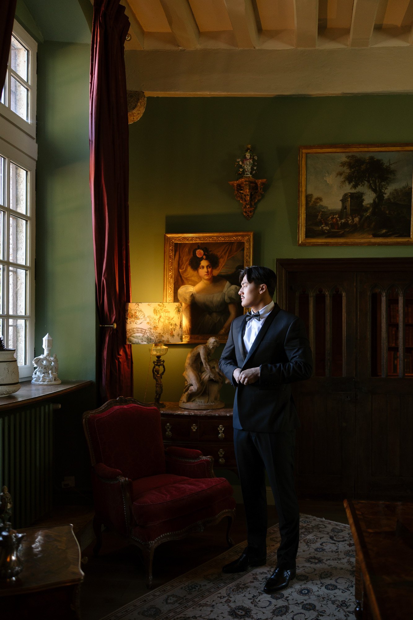 A man in a tuxedo stands in a vintage room with green walls, a large window, red curtains, classic furniture, and framed artworks. Manoir de Vacheresses wedding.