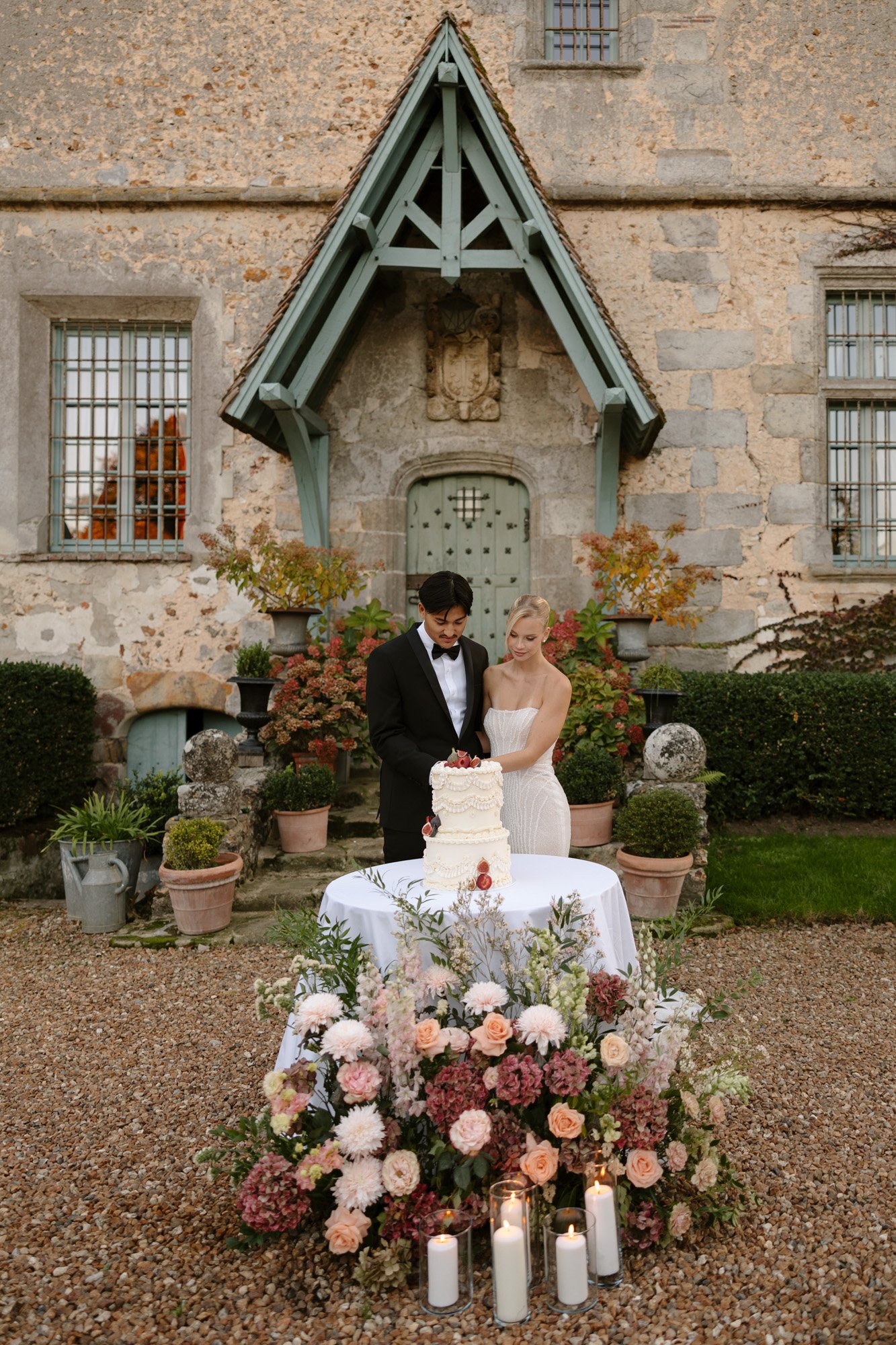 A couple stands behind a decorated wedding cake on a table with floral arrangements and candles, in front of a rustic stone building. Manoir de Vacheresses wedding.