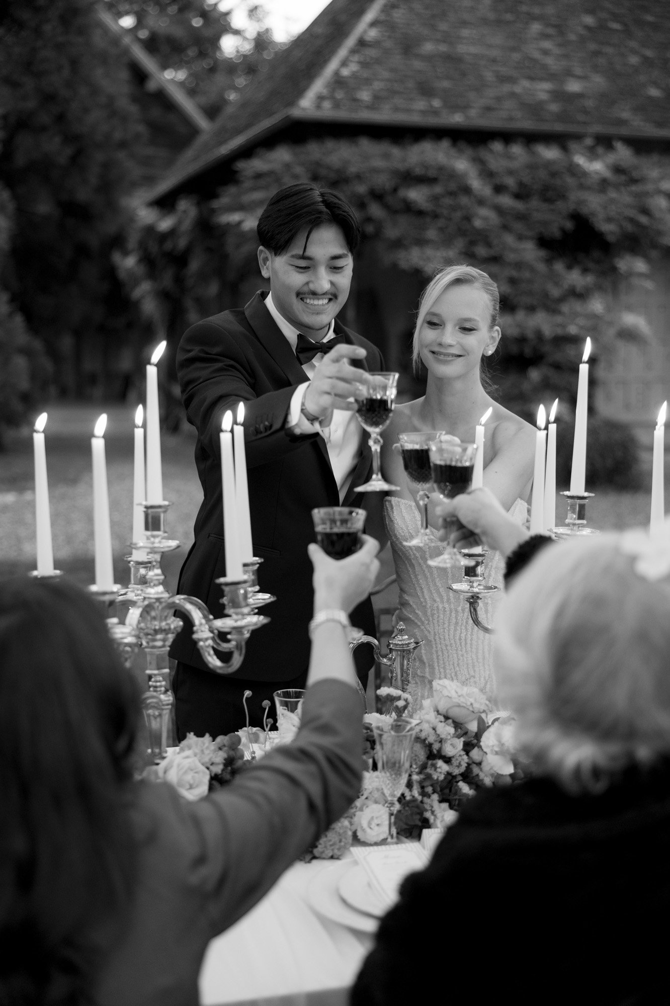 A bride and groom in formal attire stand together, smiling and raising glasses for a toast with seated guests at a candlelit outdoor table. Manoir de Vacheresses wedding.