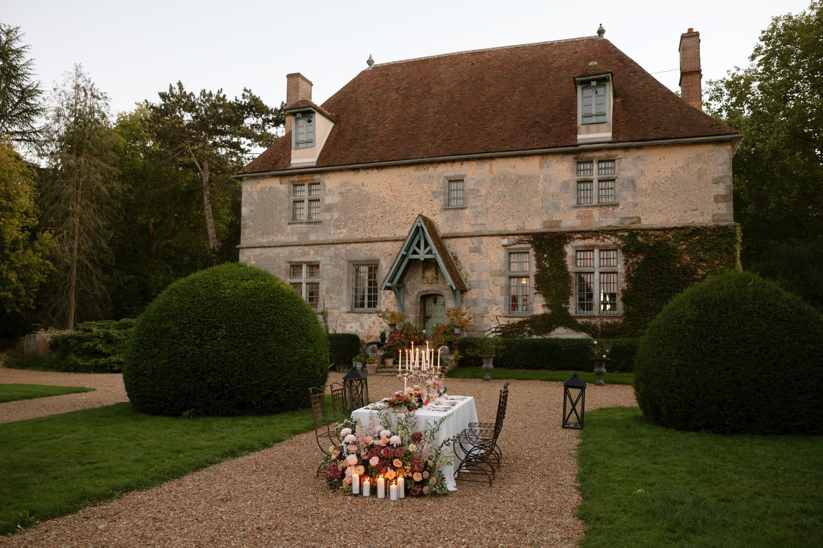 A long dining table with floral arrangements and candles is set outdoors in front of a historic stone house with a red roof and green-trimmed windows, surrounded by hedges and trees.  Manoir de Vacheresses wedding.