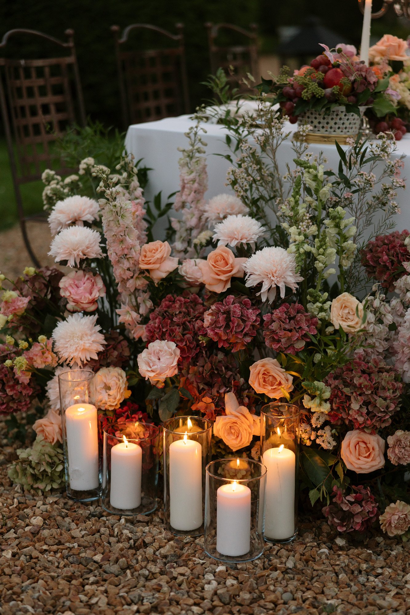 A cluster of lit white candles in glass holders sits among pink, peach, and burgundy flowers on a gravel surface next to a decorated table.