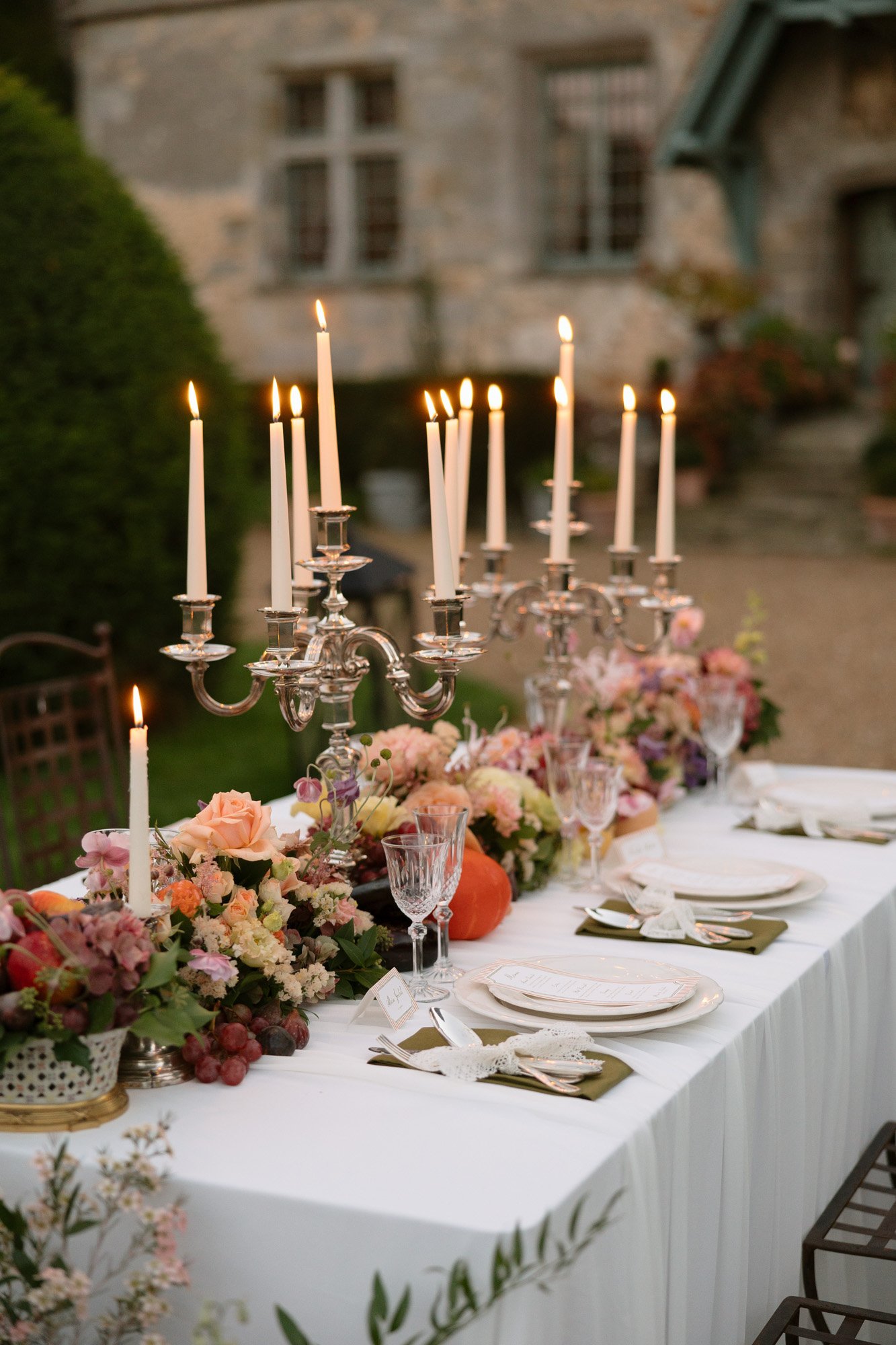 A formal outdoor dining table is set with white linens, floral arrangements, fruit, glassware, plates, and tall lit candelabras in front of a stone building.