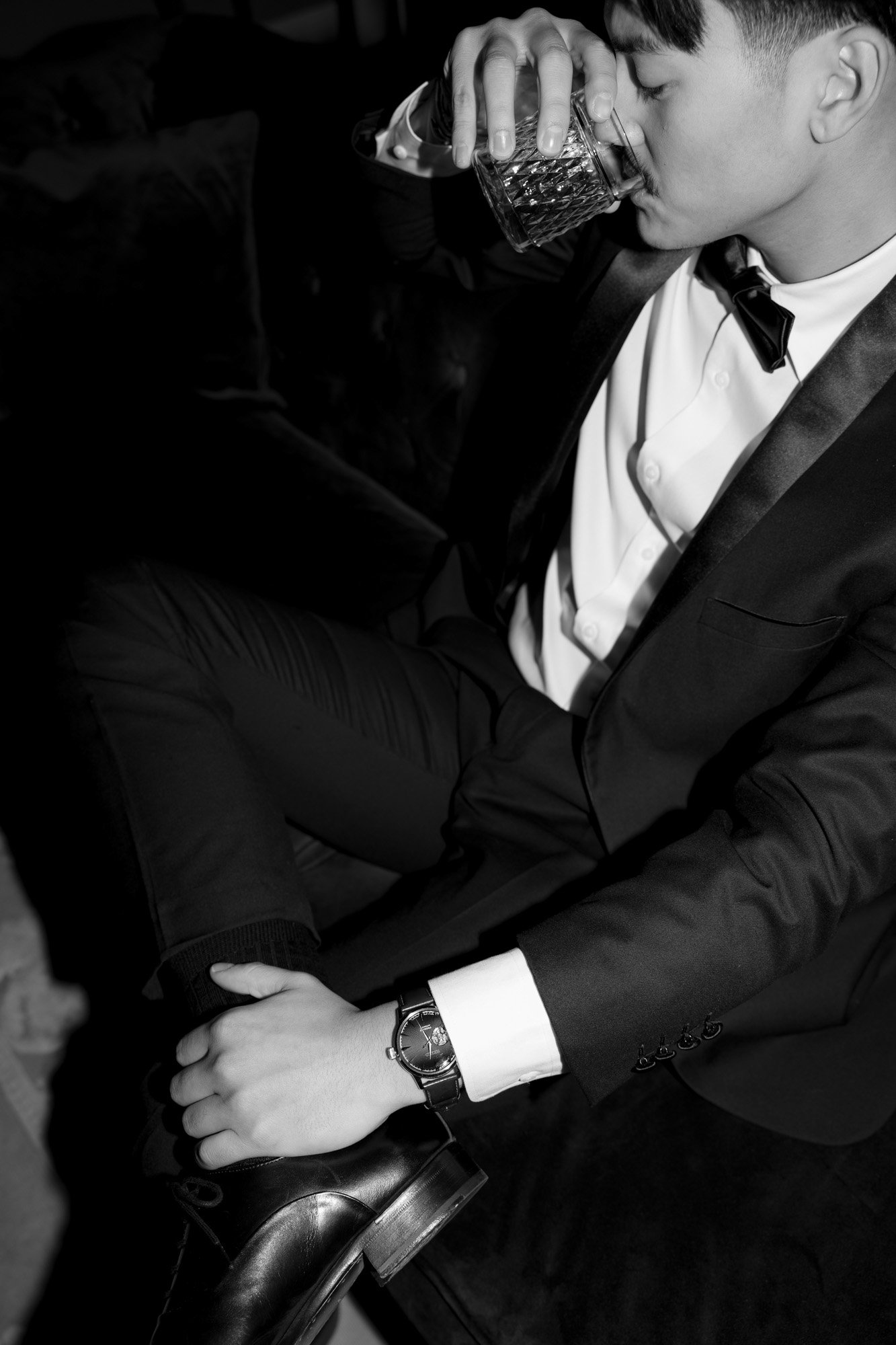 Man in a suit and bow tie sits on a couch, drinking from a glass and resting his hand on his knee.