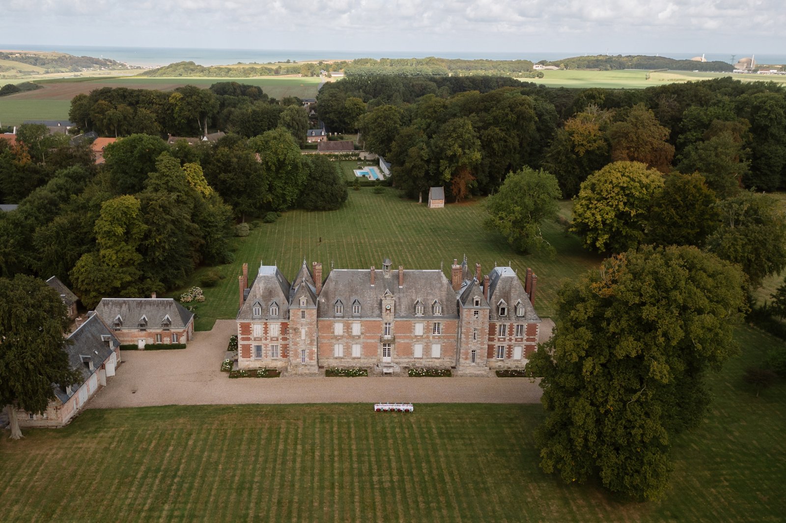 A large historic mansion with multiple chimneys stands amid expansive green lawns and surrounding trees, with countryside and sea visible in the background. Normandy Chateau Wedding.