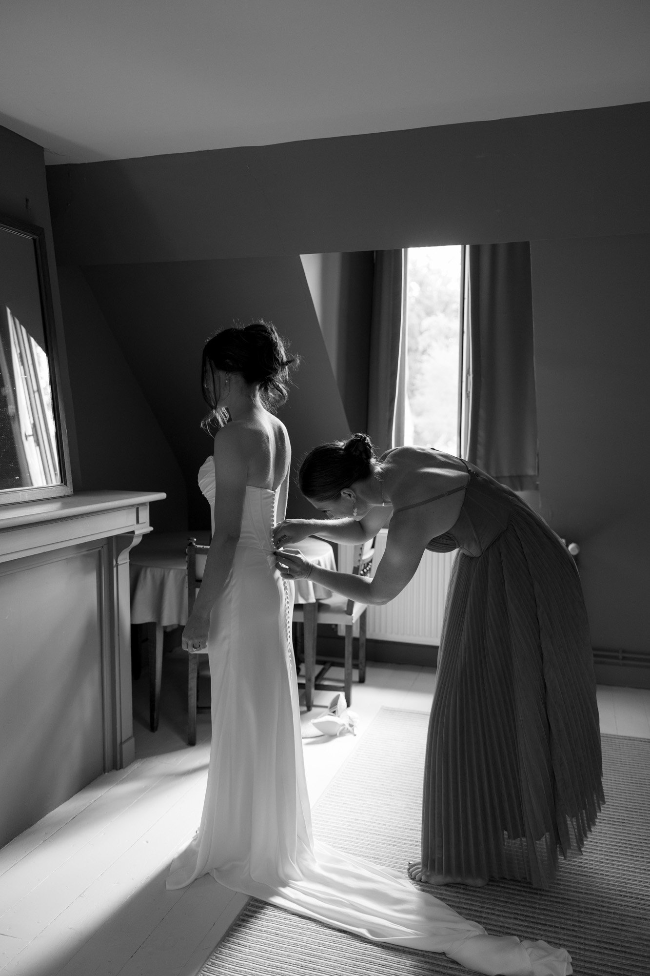 A woman in a formal dress helps another woman fasten the back of her wedding gown in a softly lit room with a window.