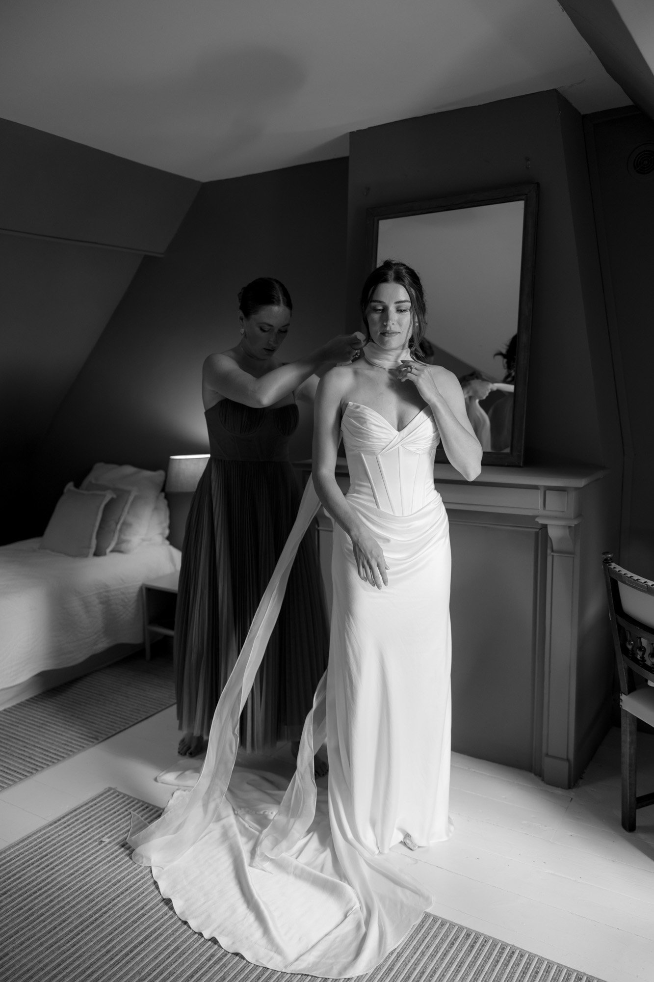 A woman in a white dress stands by a fireplace while another woman helps adjust her gown in a softly lit bedroom. Normandy Chateau Wedding.