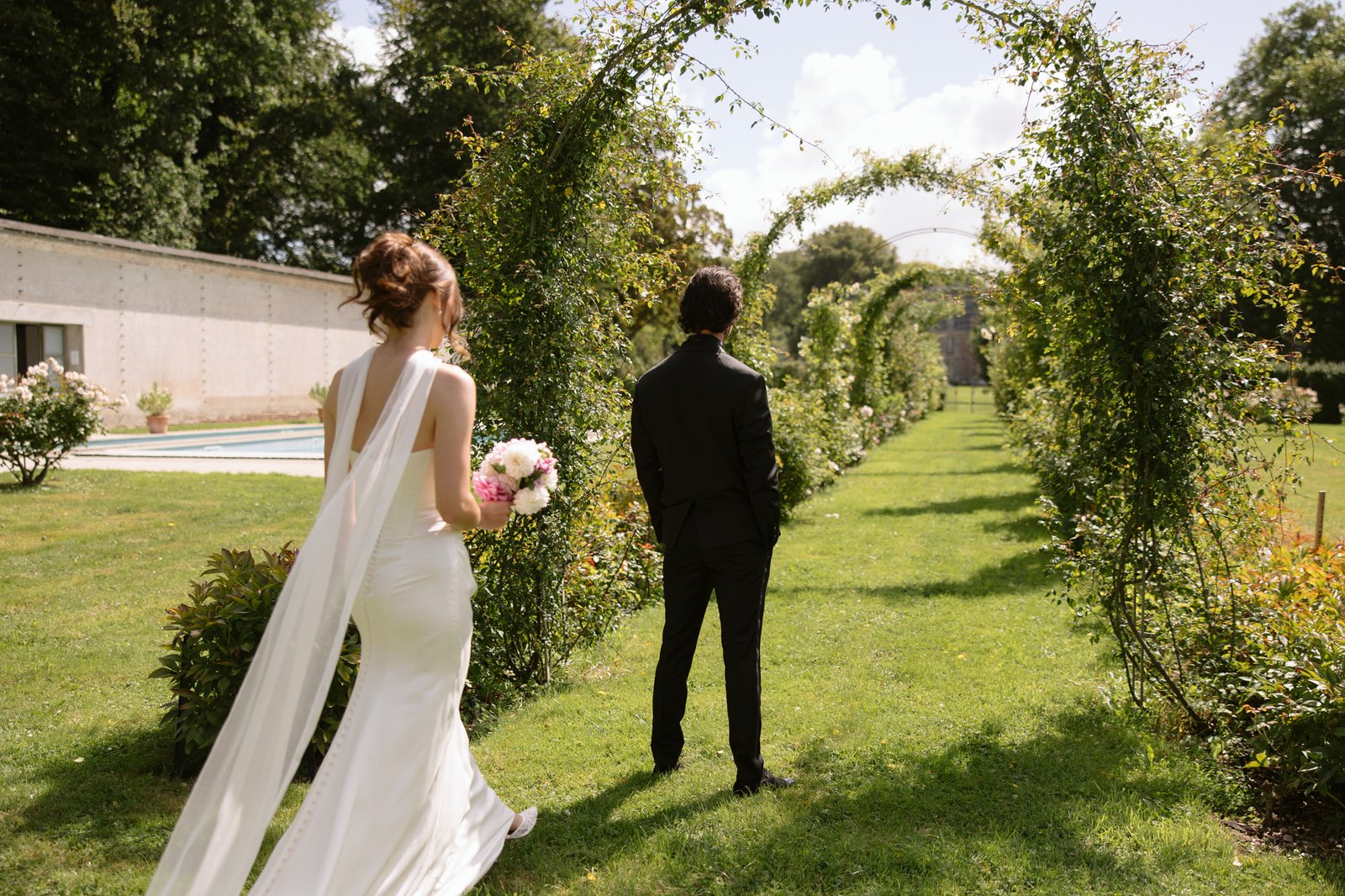 A bride in a white dress holding a bouquet walks towards a groom standing under green garden arches on a sunny day.