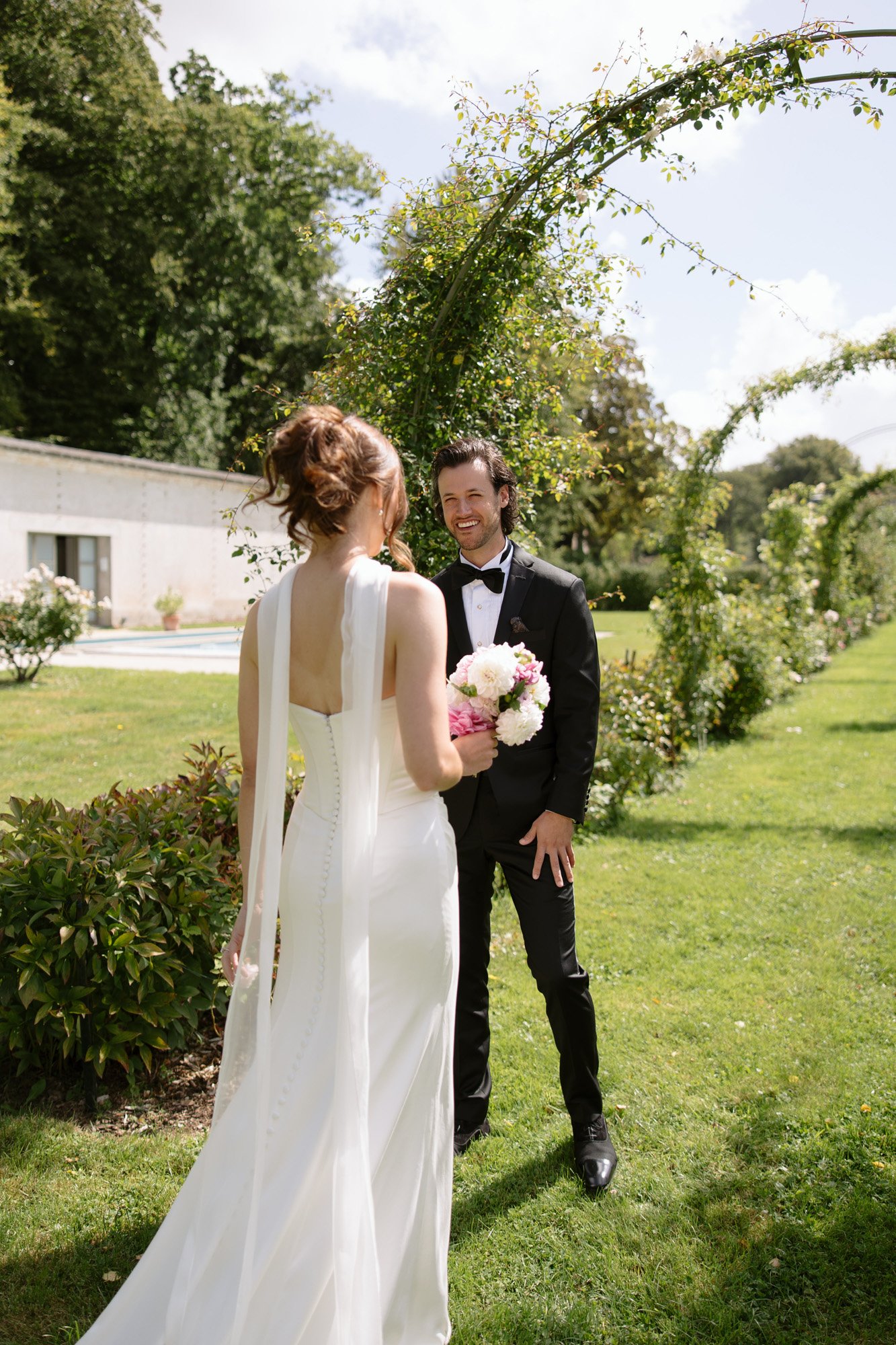 A bride in a white dress holding a bouquet stands facing a smiling groom in a black tuxedo outdoors on a green lawn with arching greenery.
