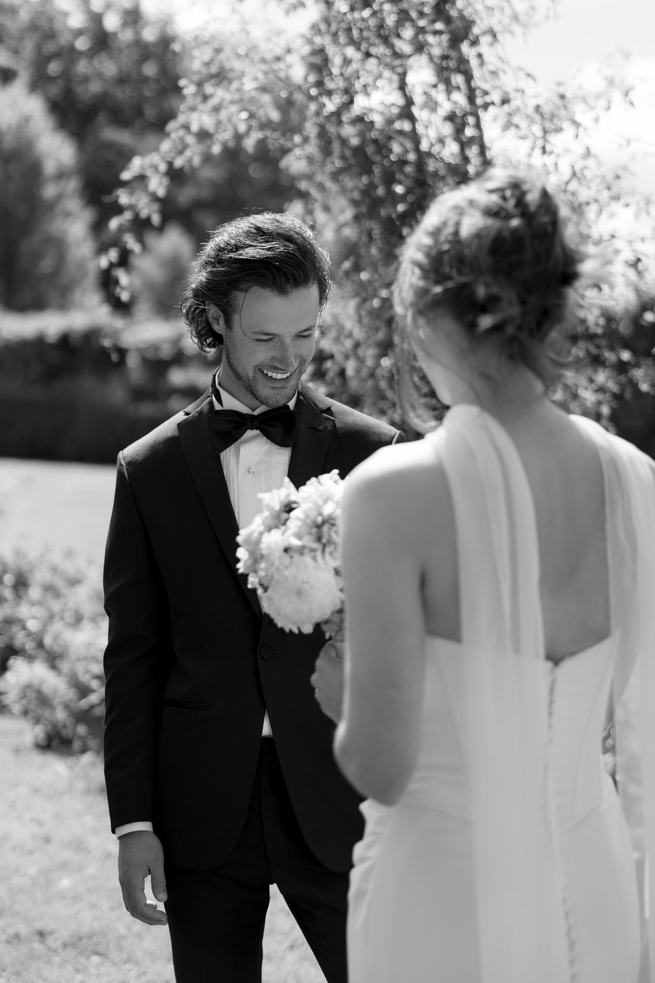 A man in a tuxedo and a woman in a wedding dress stand outdoors, facing each other. The woman holds a bouquet. The scene is bright and surrounded by greenery.