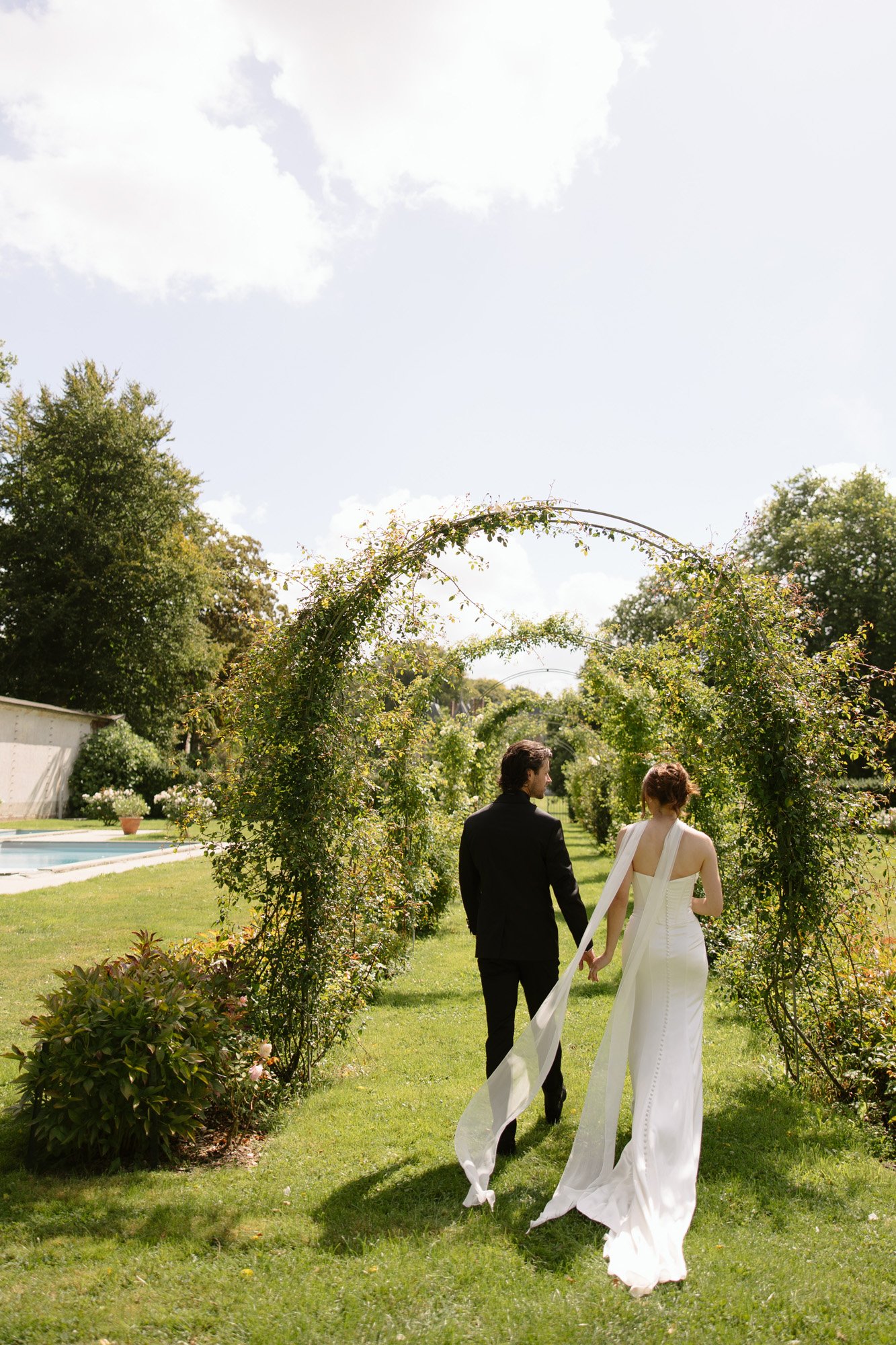 A couple in formal attire walks hand in hand through a garden archway, with greenery and a pool visible in the background.