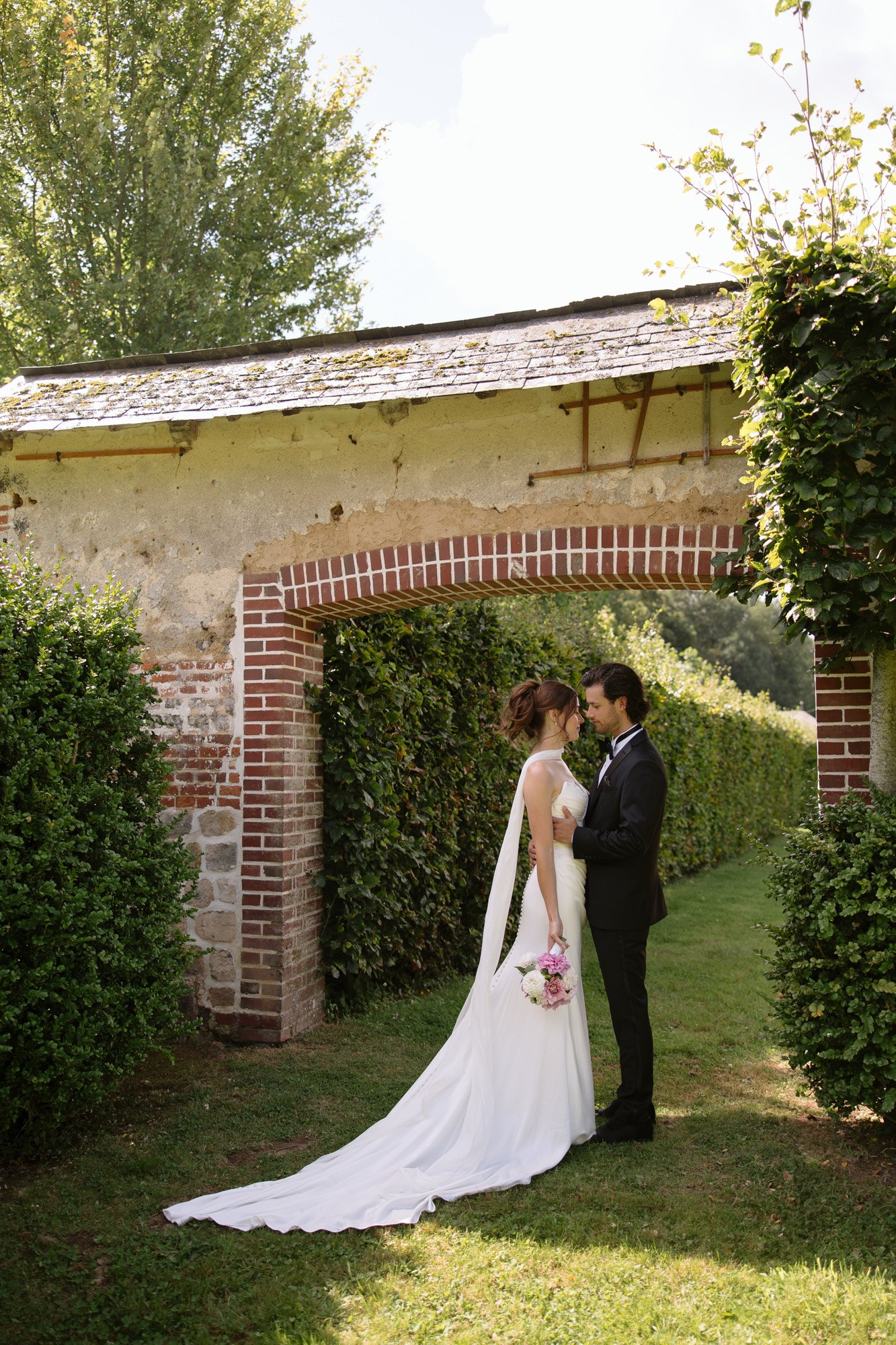 A bride and groom stand facing each other under a brick archway in a garden, with greenery and sunlight surrounding them. The bride holds a bouquet and wears a long white dress and veil.