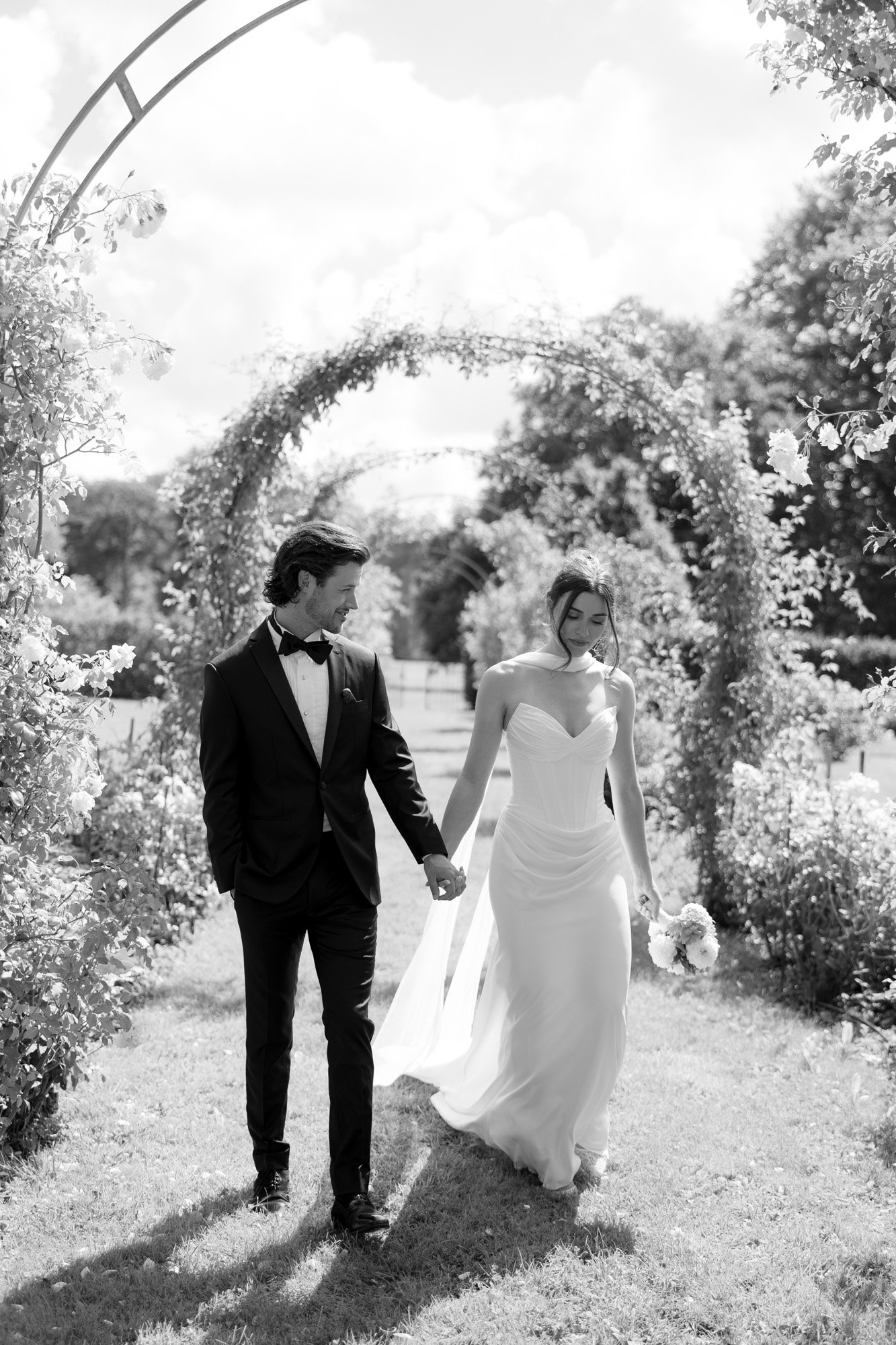 A bride and groom, dressed in wedding attire, walk hand in hand through a garden archway on a sunny day.