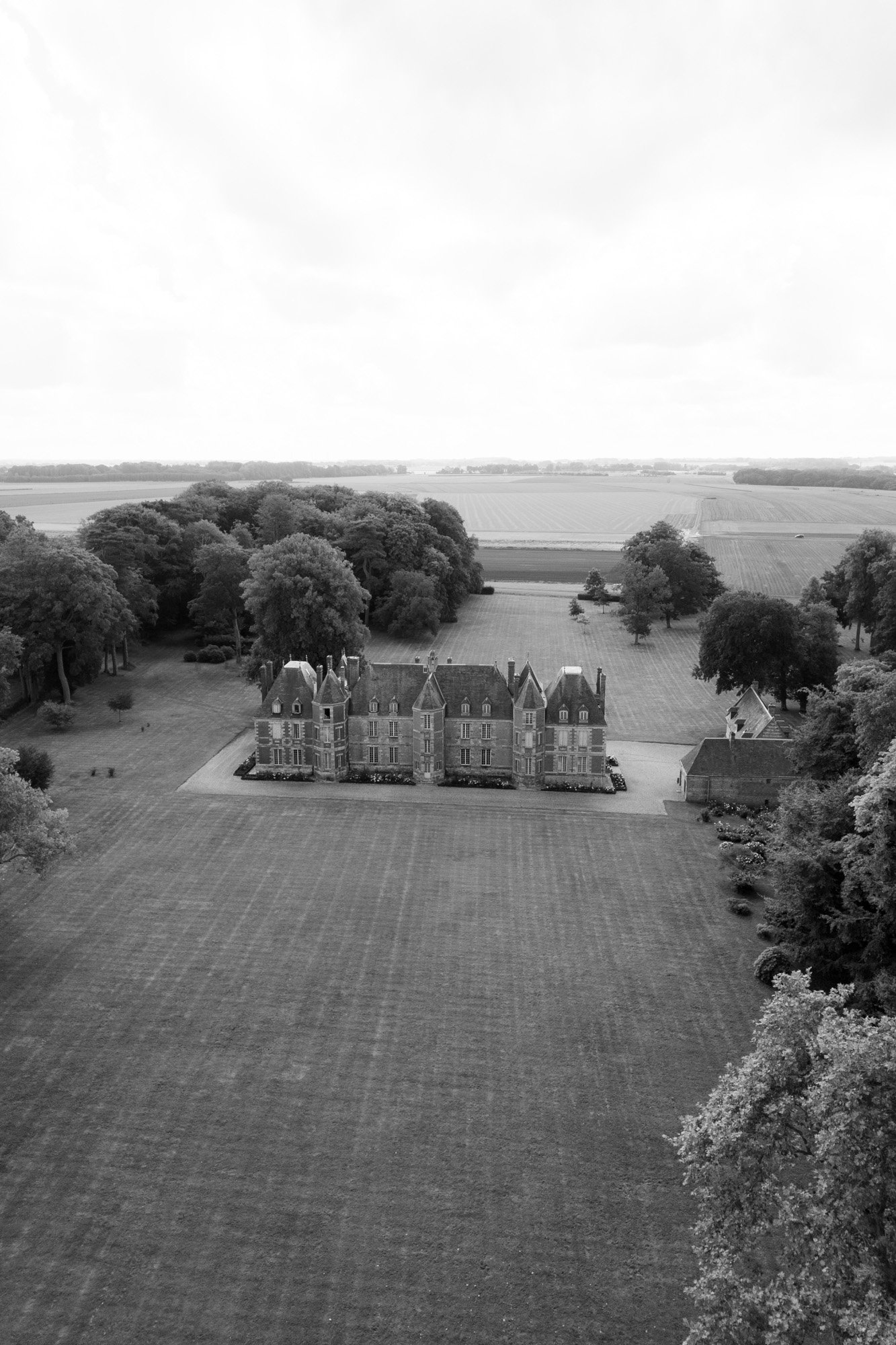 A large manor house with peaked roofs sits centered on a vast, neatly mowed lawn, surrounded by trees and open fields under a cloudy sky.