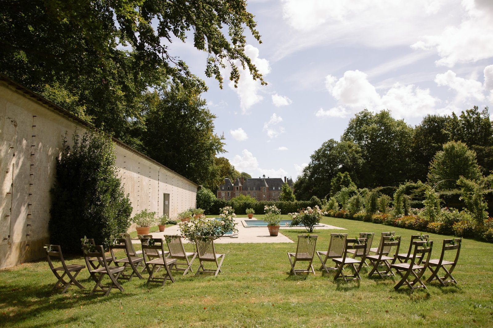 Wooden chairs are arranged in rows on a lawn facing a pool, with a stone building on the left and lush gardens in the background under a partly cloudy sky.