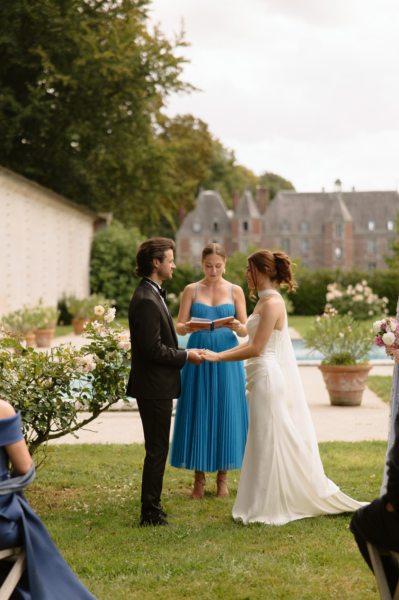 A bride and groom hold hands during an outdoor wedding ceremony, with an officiant standing between them. A large building and garden are visible in the background. Normandy Chateau Wedding.