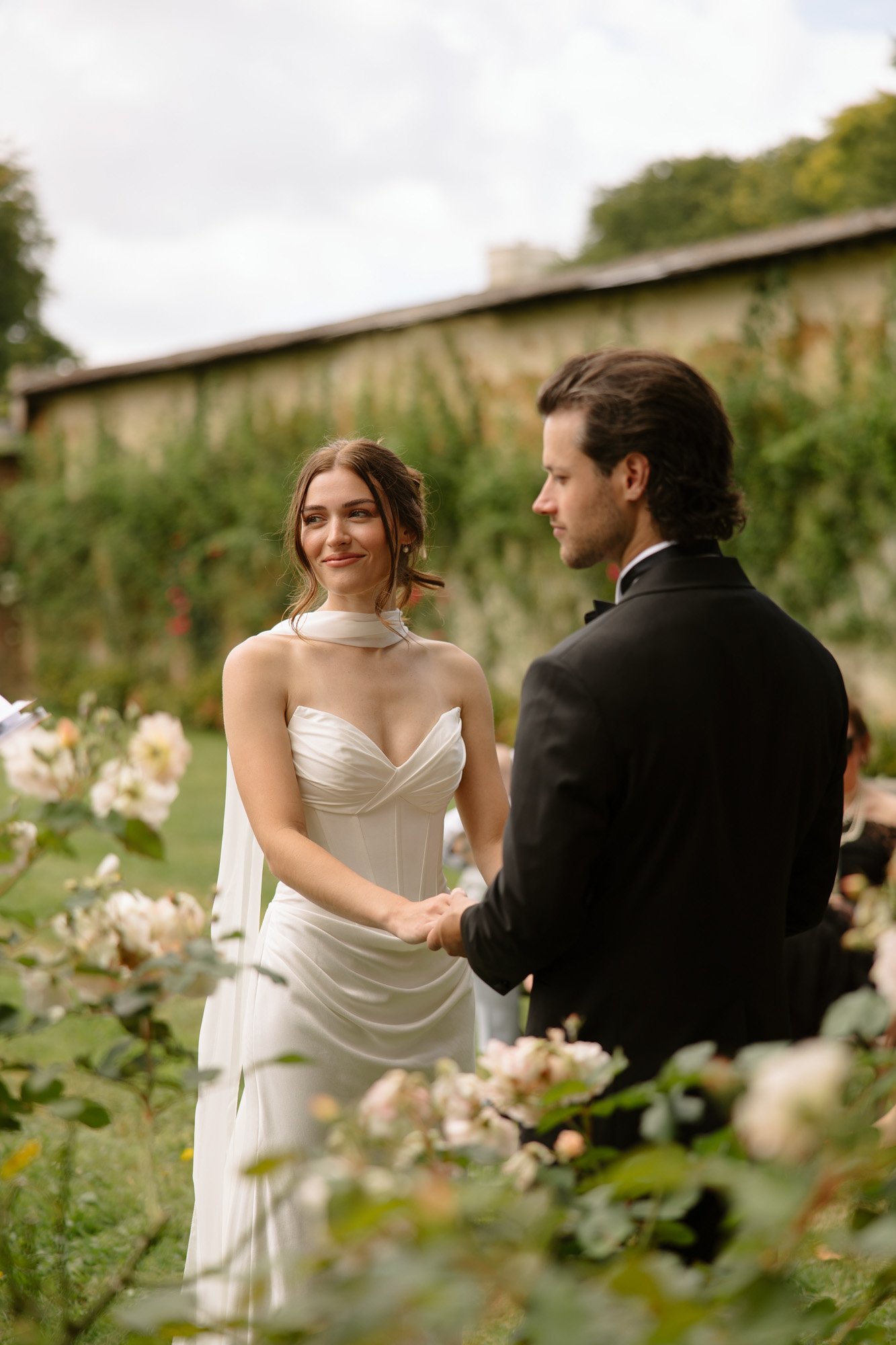 A bride and groom stand facing each other holding hands outdoors, surrounded by flowers, during a wedding ceremony. Normandy Chateau Wedding.