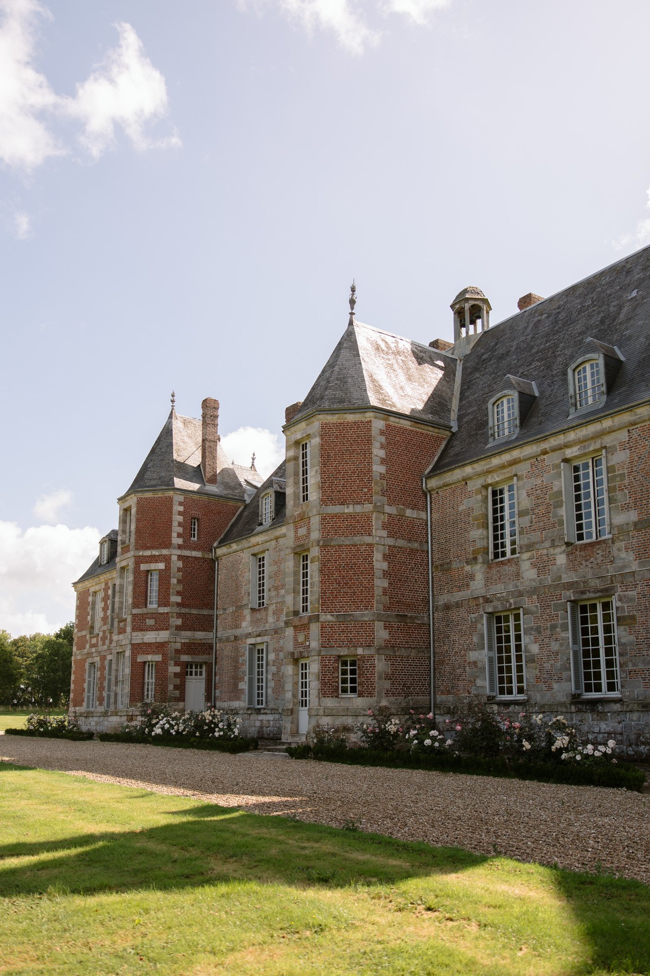 A large historic brick and stone building with multiple windows and steep slate roofs, surrounded by a gravel path and green lawn under a partly cloudy sky. Normandy Chateau Wedding.