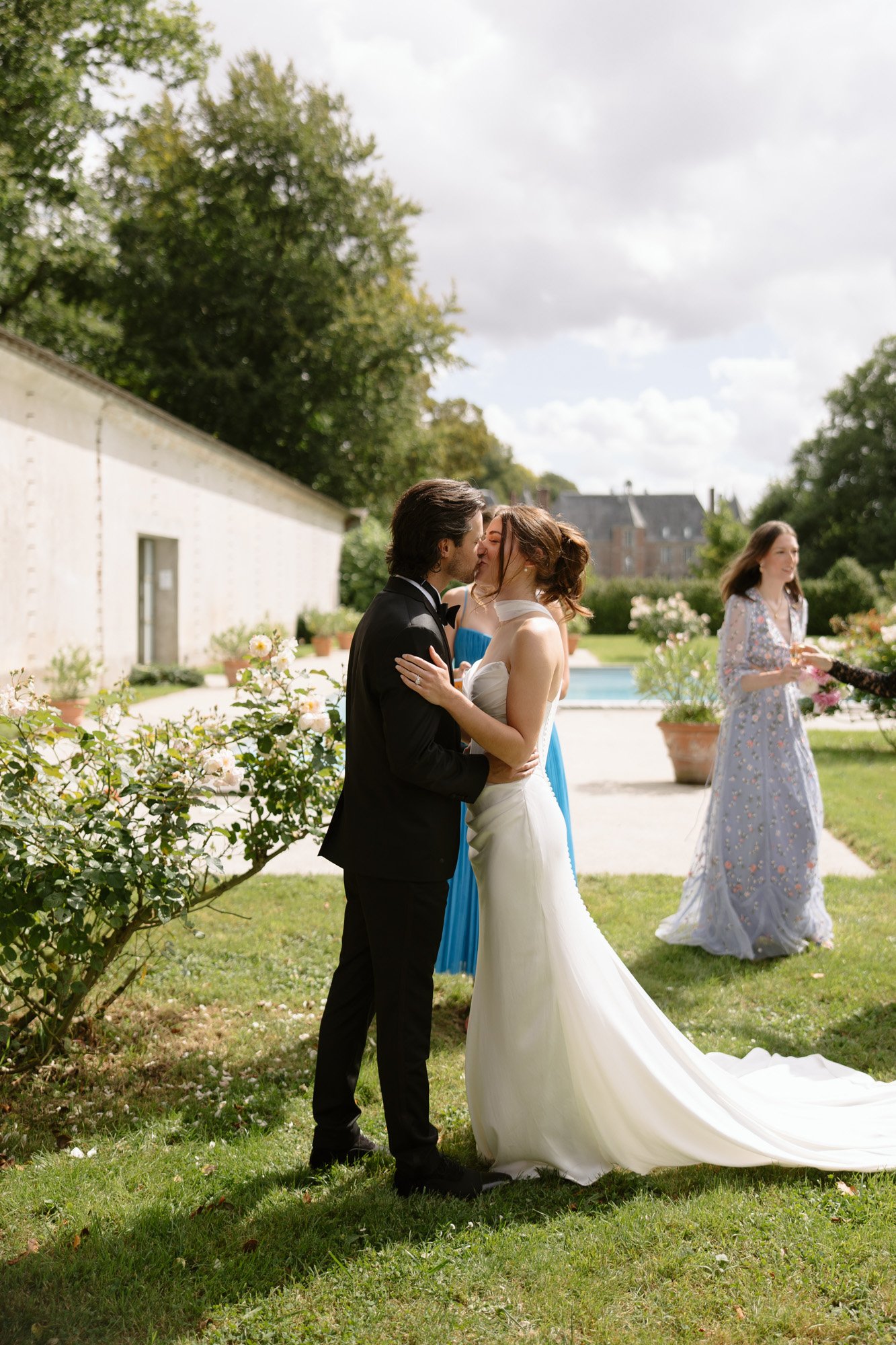 A bride and groom kiss outdoors on a lawn near a pool, while two women in formal dresses stand and walk in the background.
