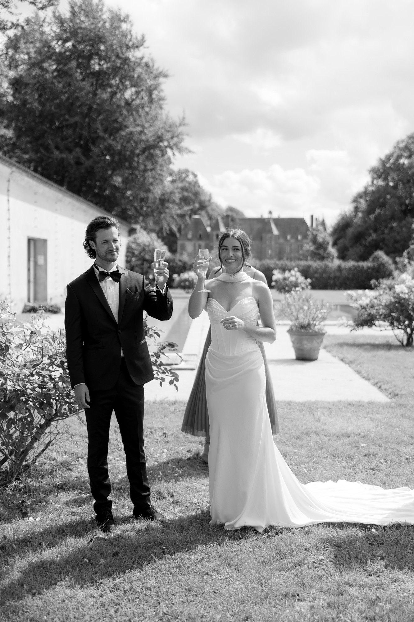 A man in a suit and a woman in a wedding dress stand on grass, holding up glasses and smiling outside on a sunny day.