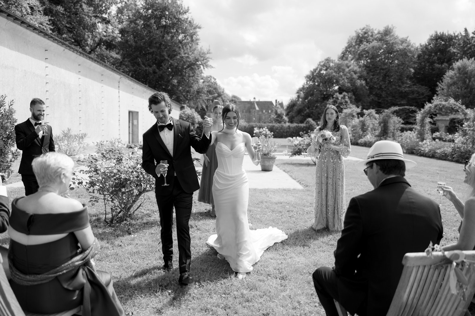 A bride and groom walk hand in hand outdoors, surrounded by formally dressed guests seated and standing on a lawn during a wedding celebration. Normandy Chateau Wedding.